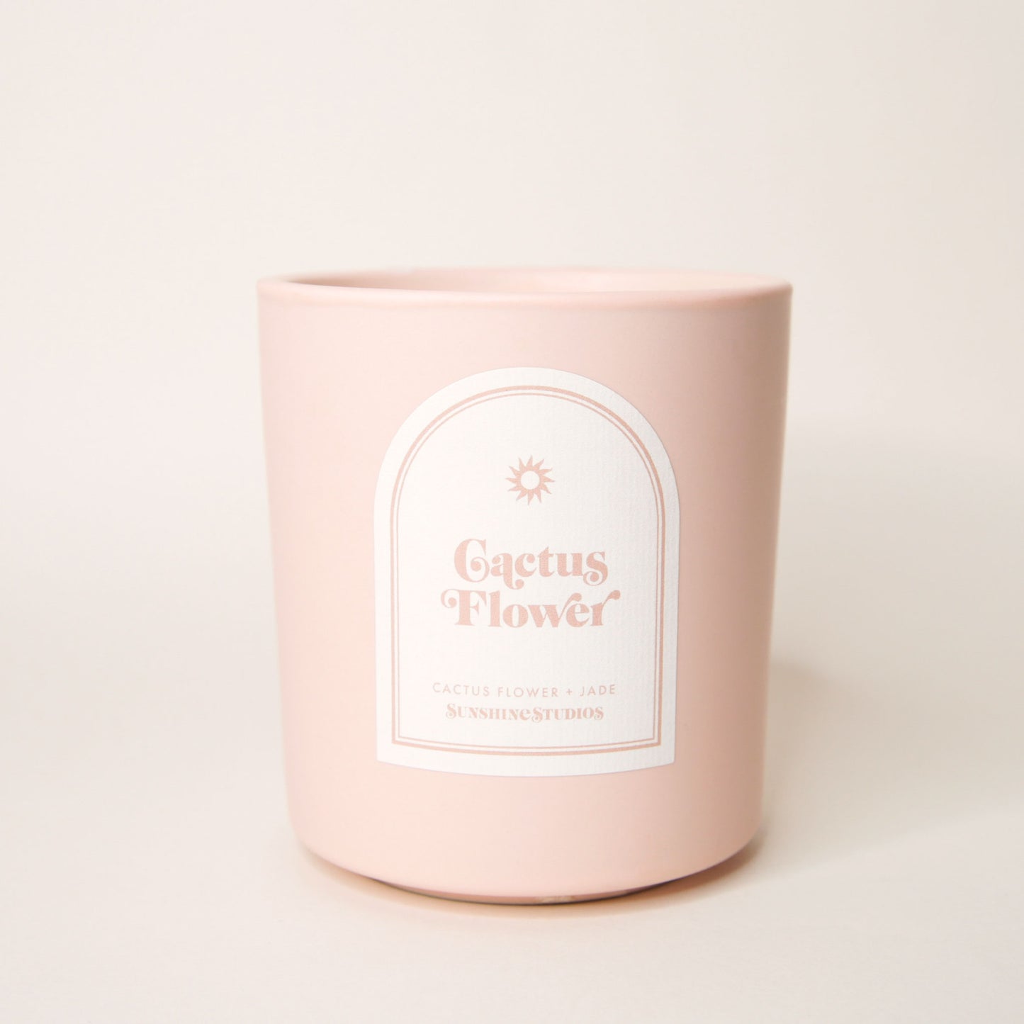 Pastel pink candle with an arched label that reads 'Cactus Flower, cactus flower + jade, sunshine studios' in soft pink lettering. A small sun design sits above the text.