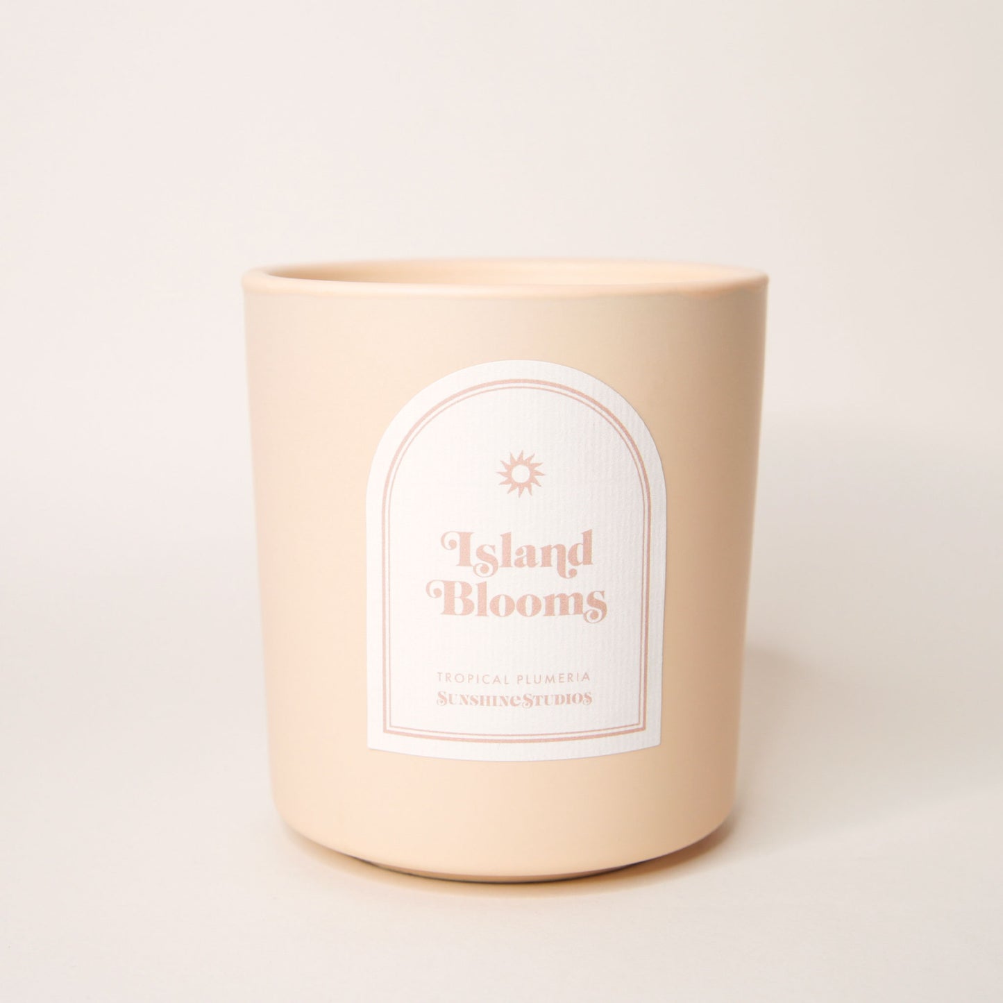 Soft beige candle with an arched label that reads 'Island Blooms, tropical plumeria, sunshine studios' in soft pink lettering. A small sun design sits above the text.
