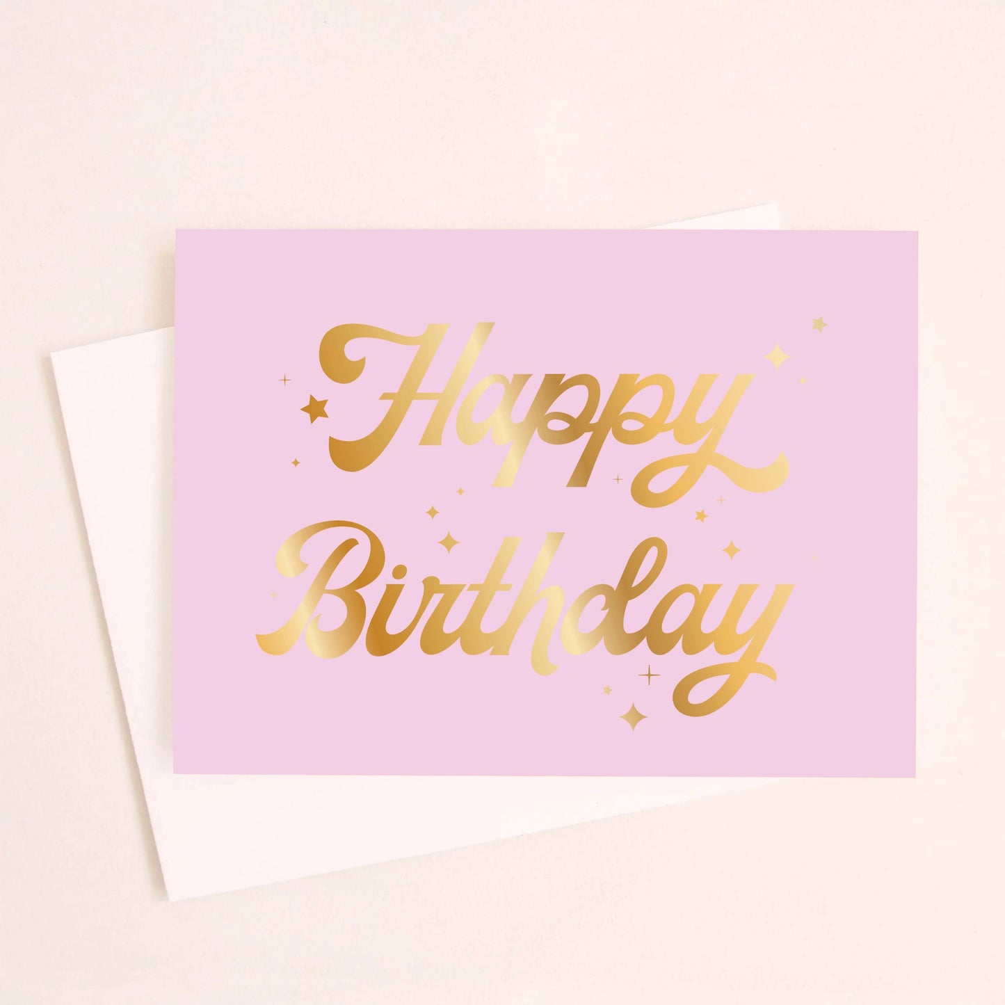 On a cream background is a lavender card with gold foiled cursive text that reads, "Happy Birthday" along with a white envelope.