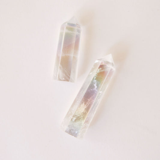 A photo of two aura clear quartz crystals, translucent with white accents and rainbow hues on the inside. The two sizes represent the two sizes available for purchase. Each crystal is sold separately. These aura clear quartz crystals are in the shape of a point and are flat on the bottom to allow them to be freestanding.
