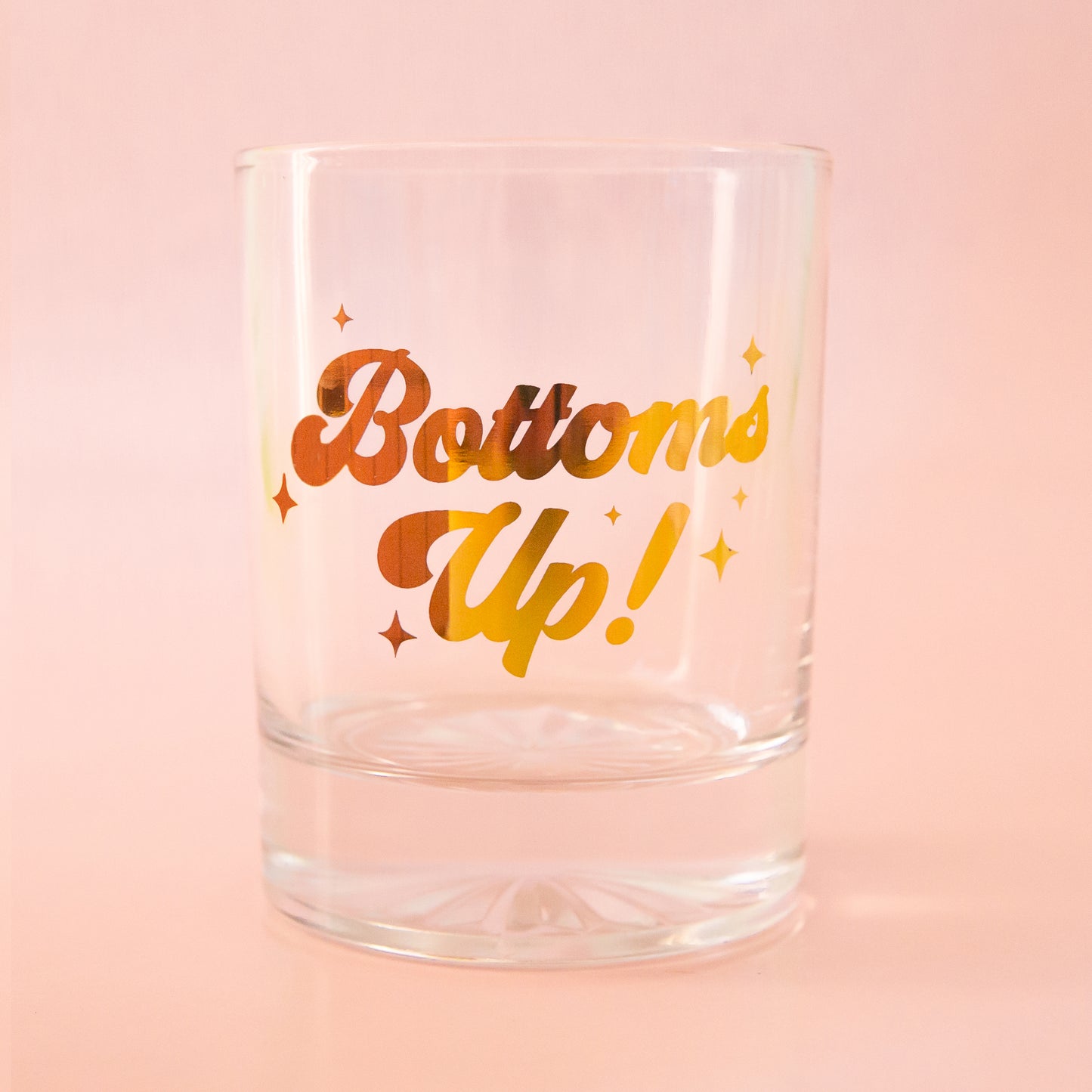 Glass tumbler with gold foil writing, spelling"bottoms up!" is shown against a light pink background