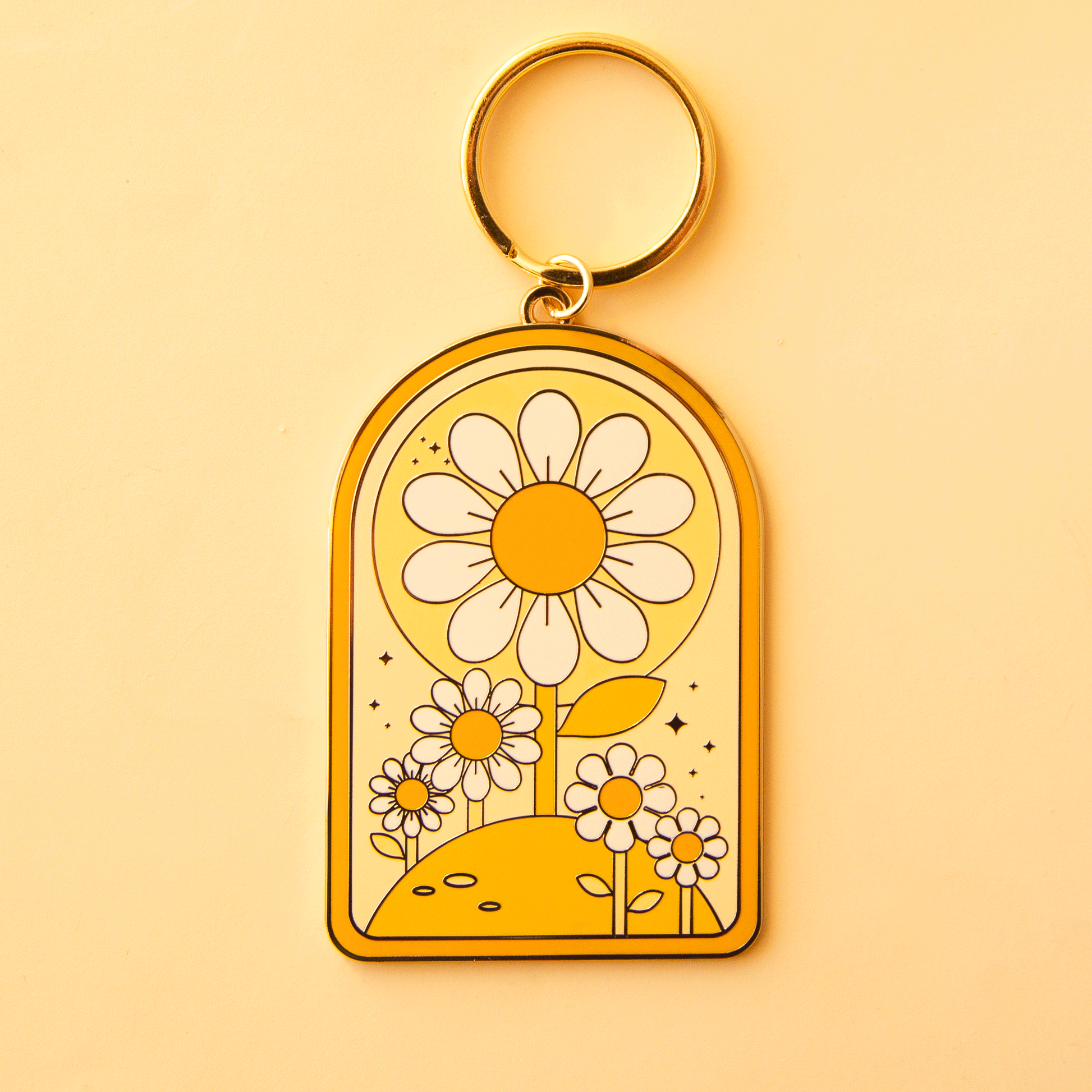 On a yellow background is an arched keychain with a gold loop and a white and yellow daisy design.