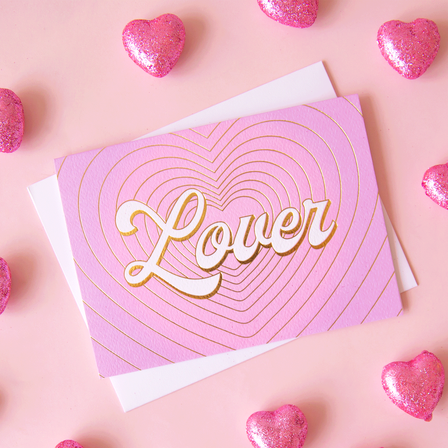 On a pink background is a pink and purple gradient card with gold foiled heart shaped outlines and ivory text that reads, "Lover".