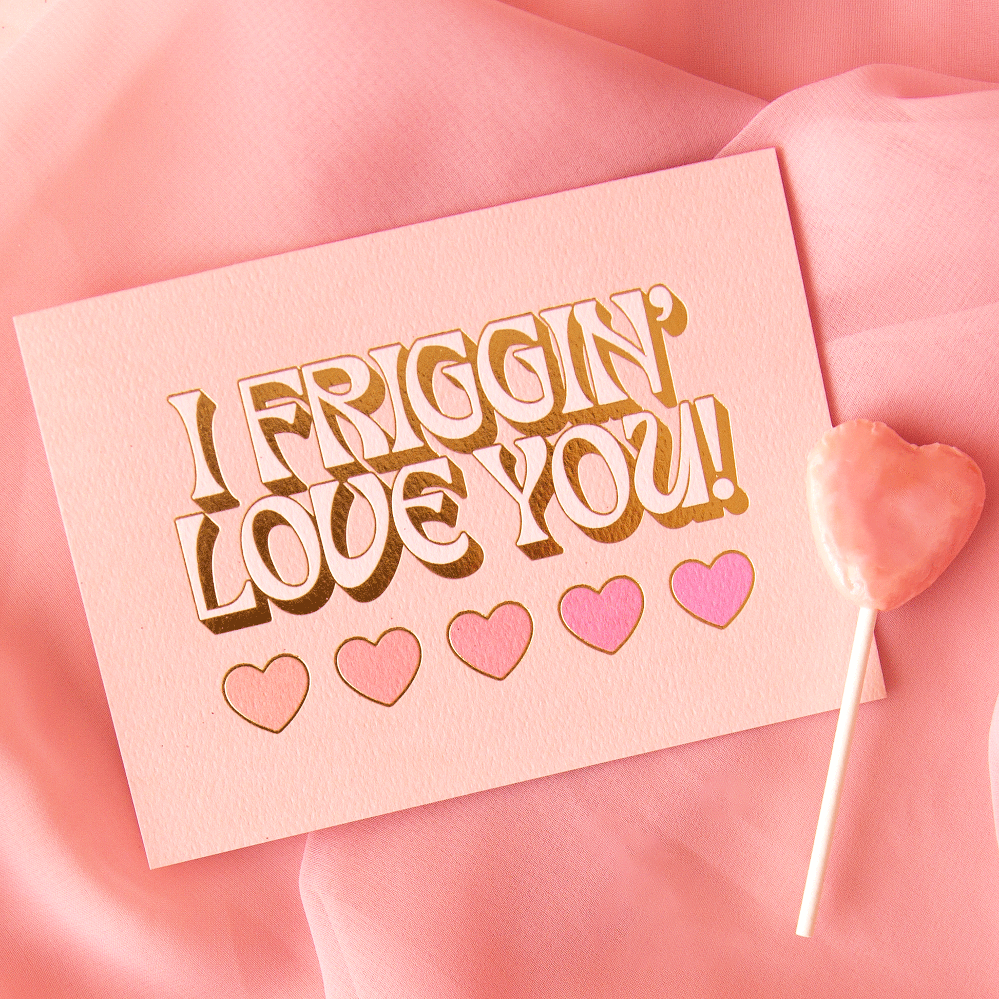 On a pink background is a light pink card with text that reads, "I Friggin' Love You!" with five hearts underneath and staged next to a heart shaped sucker that is not included with purchase.