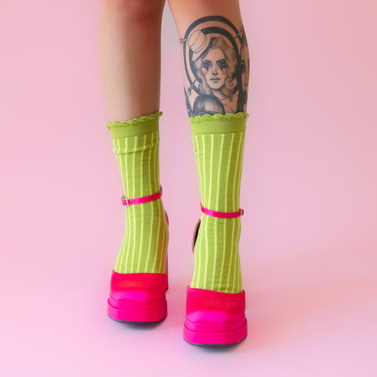 A pair of lime green crew socks with a lighter green stripe and a ruffle edge at the top. Worn on a model with hot pink satin closed toe high heels. 