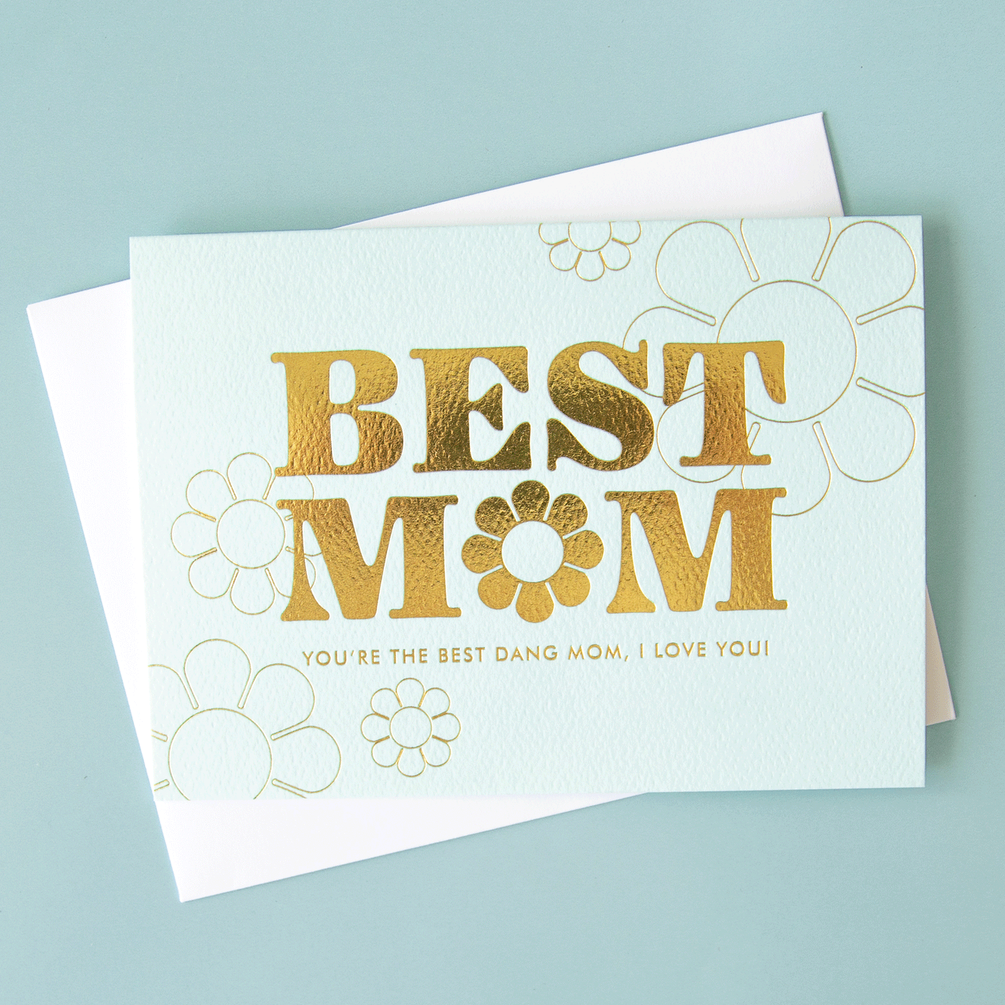 On a blue background is a light blue card with gold foiled text that reads, "Best Mom You're The Best Dang Mom, I Love You" along with a flower shape in the place of the 'O' on 'Mom'.