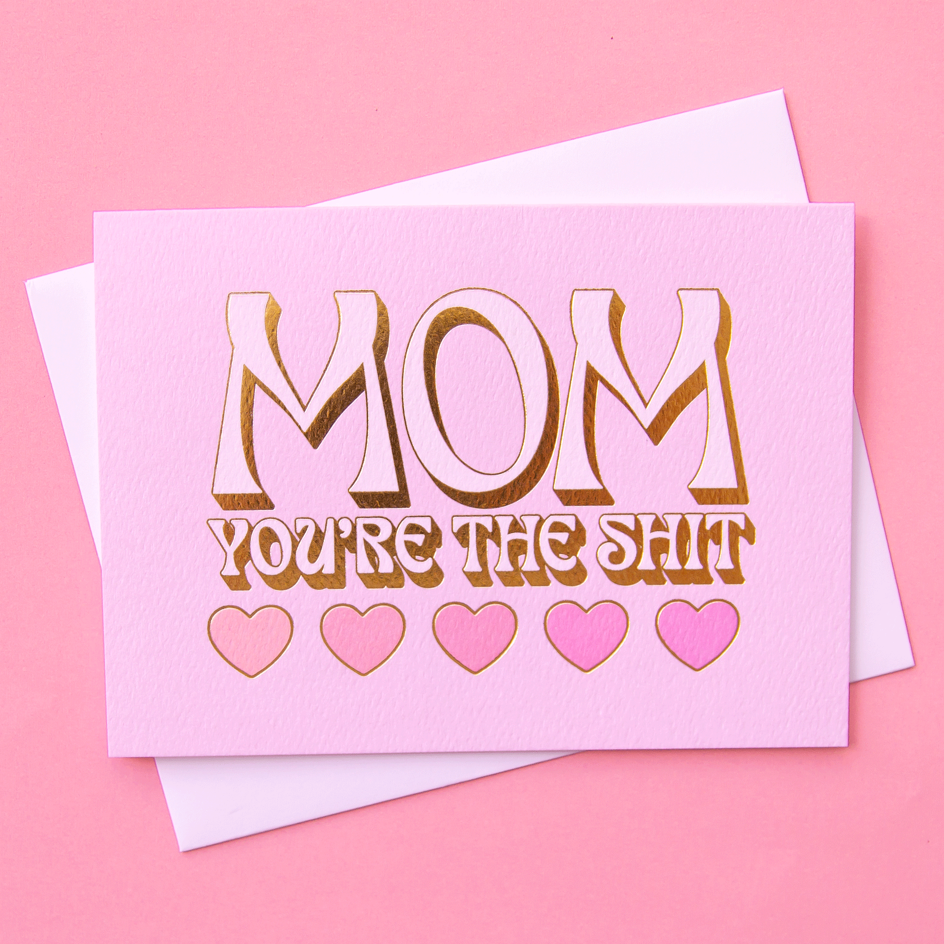 On a pink background is a pink card with heart shapes and text above that reads, "Mom You're The Shit".