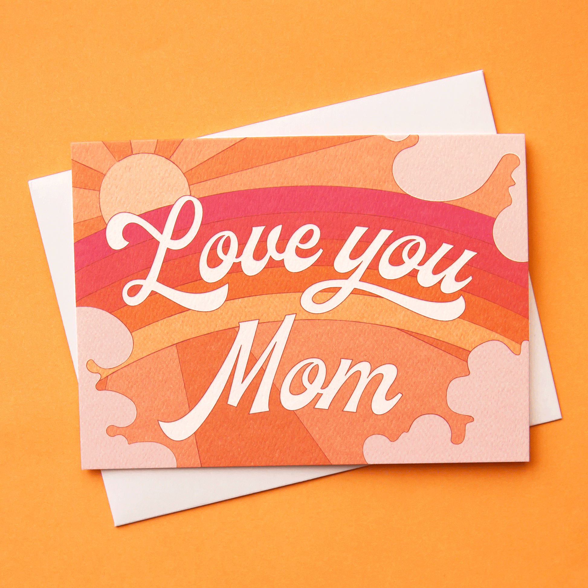 A card with a rainbow graphic made with shades or orange, pink and red along with a shining sun, subtle clouds around the edge and white cursive text that reads, "Love you Mom" in the center as well as a white envelope.