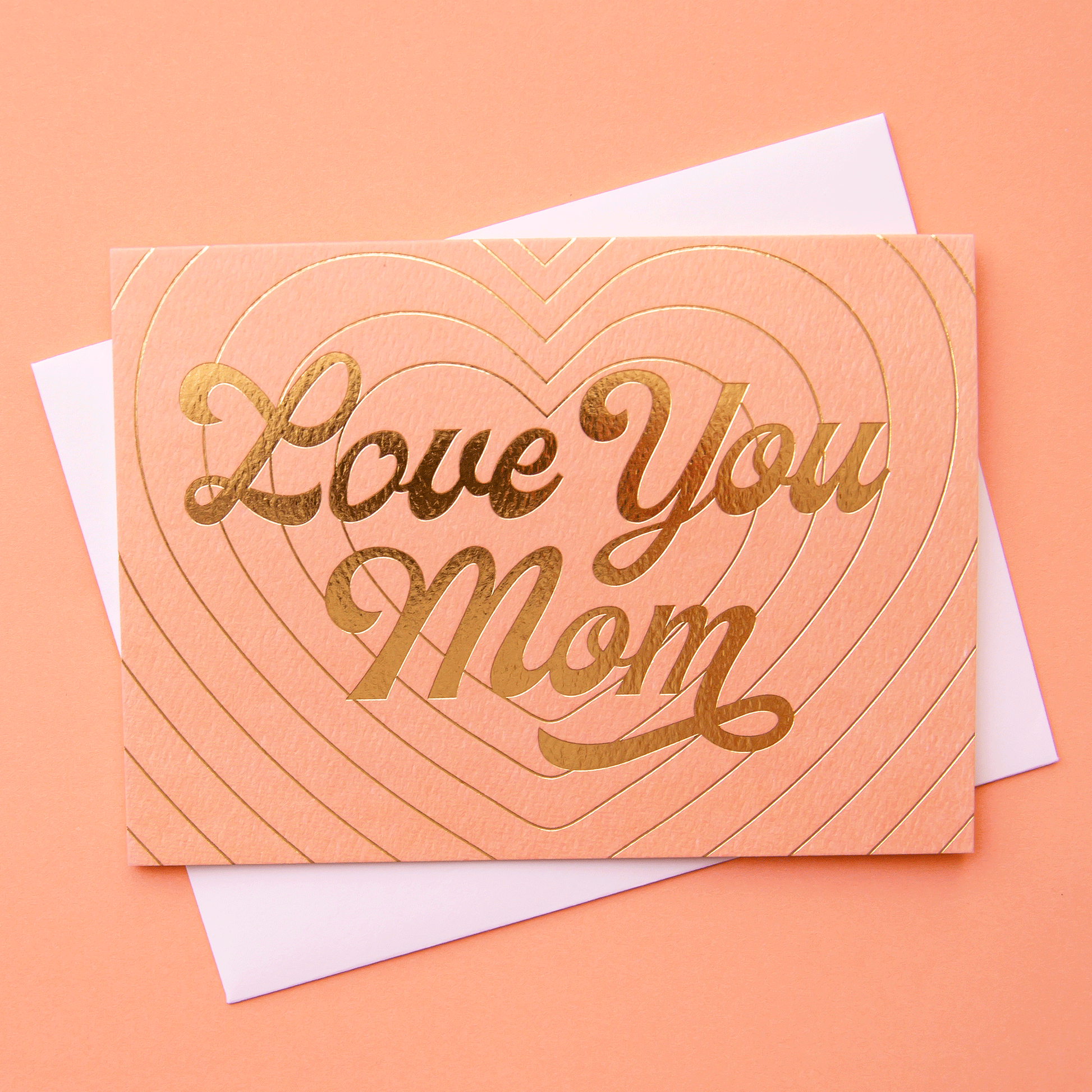 Peach card that reads 'love you mom' in gold foil. The card is filled with layers of a beaming gold foil heart design behind and is accompanied by with a white envelope.