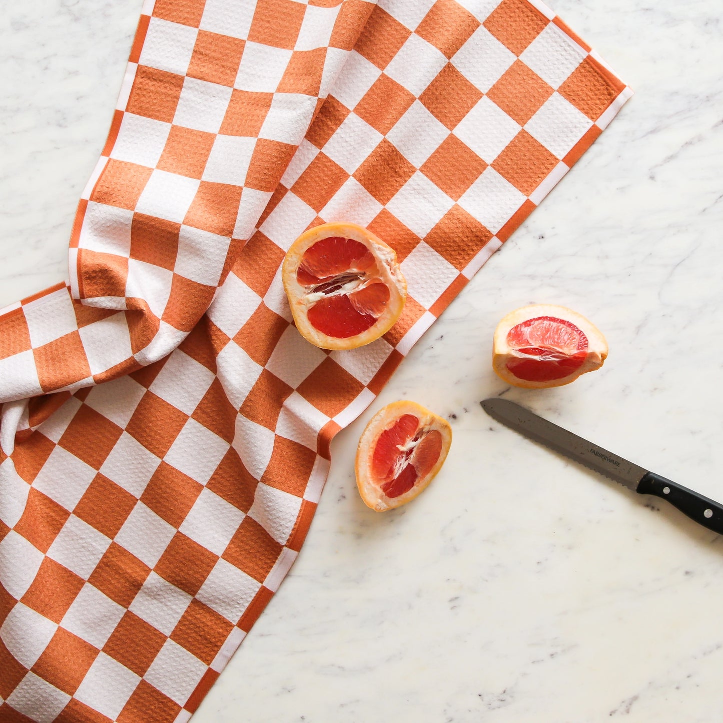 On a marble backdrop is an orange and ivory checkered kitchen towel with a staged blood orange cut up next to it.