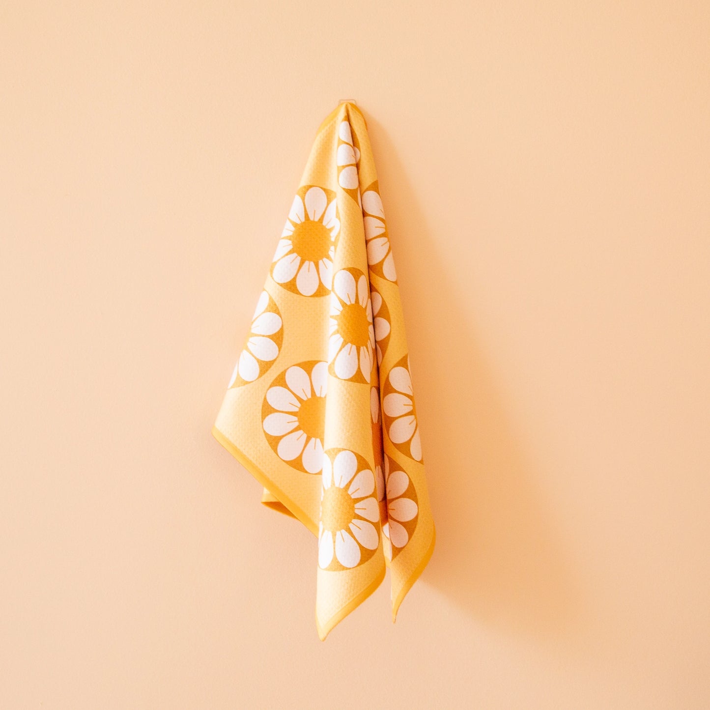 On a peachy background is a yellow and white daisy kitchen towel with a waffle texture and hung up on a hook.