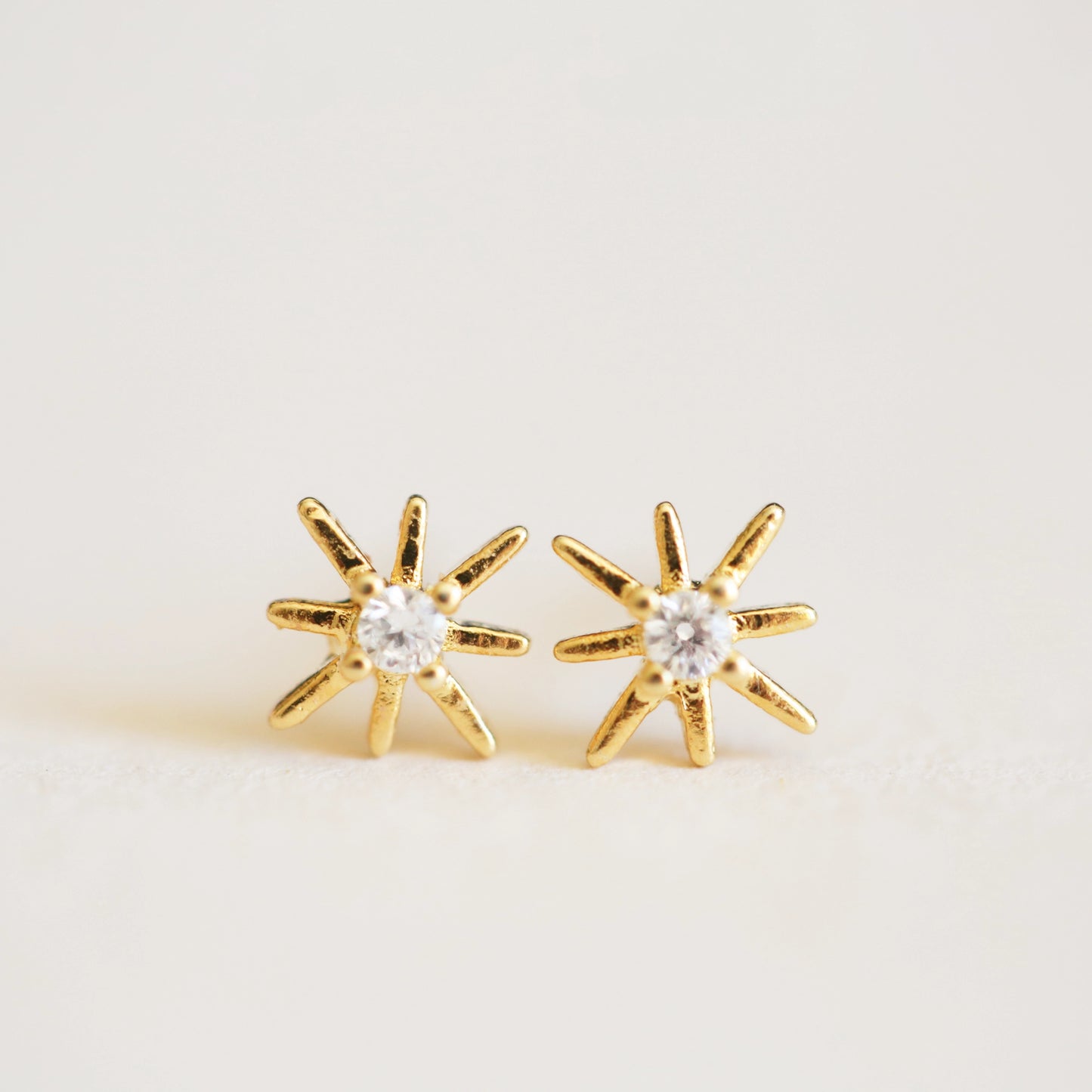 On an ivory background is a pair of dainty gold star earrings with a CZ stone in the center. 
