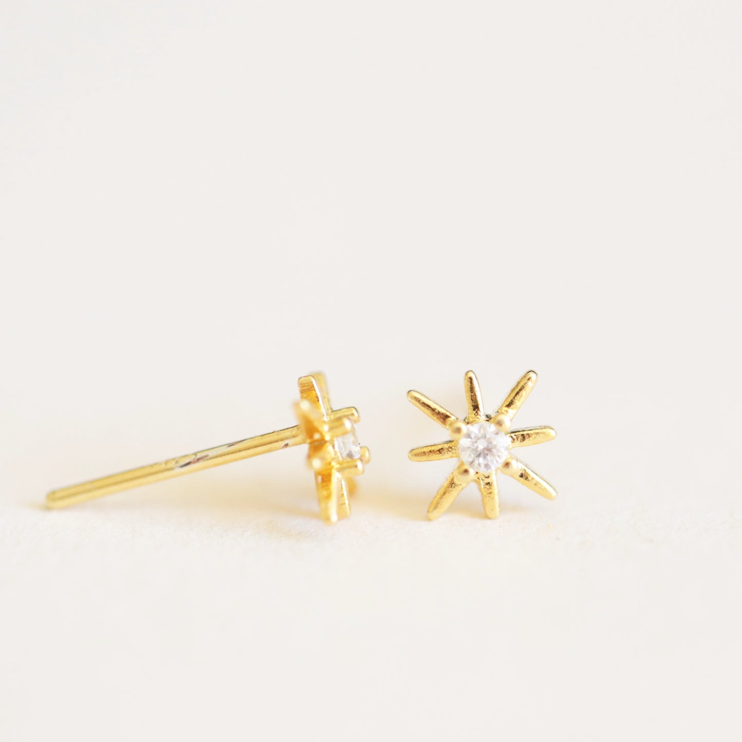 On an ivory background is a pair of dainty gold star earrings with a CZ stone in the center.