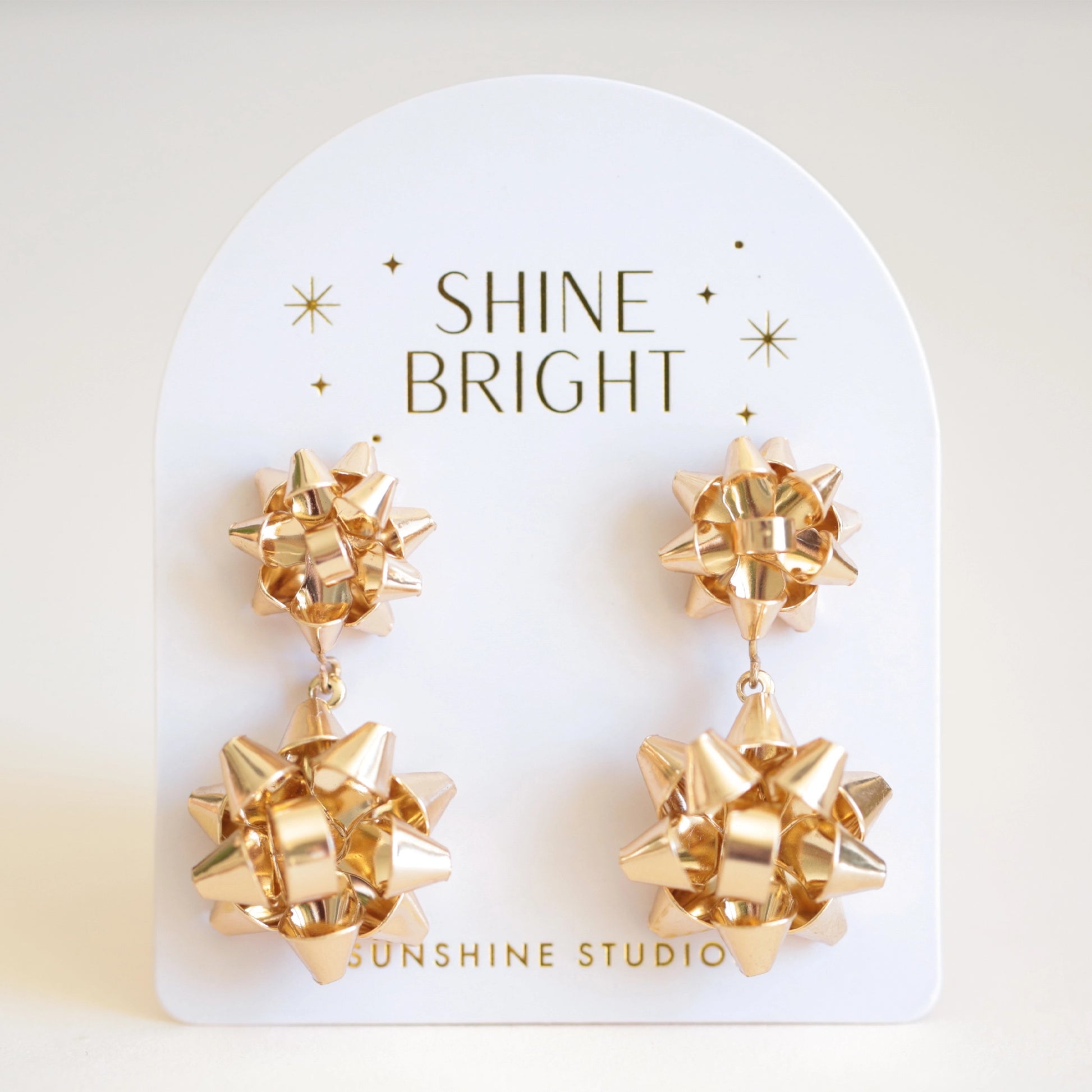 On an ivory background is a pair of gold holiday bow shaped earrings that have a smaller gold bow and a dangling larger bow attached to the bottom. The packaging reads, "Shine Bright Sunshine Studios". 