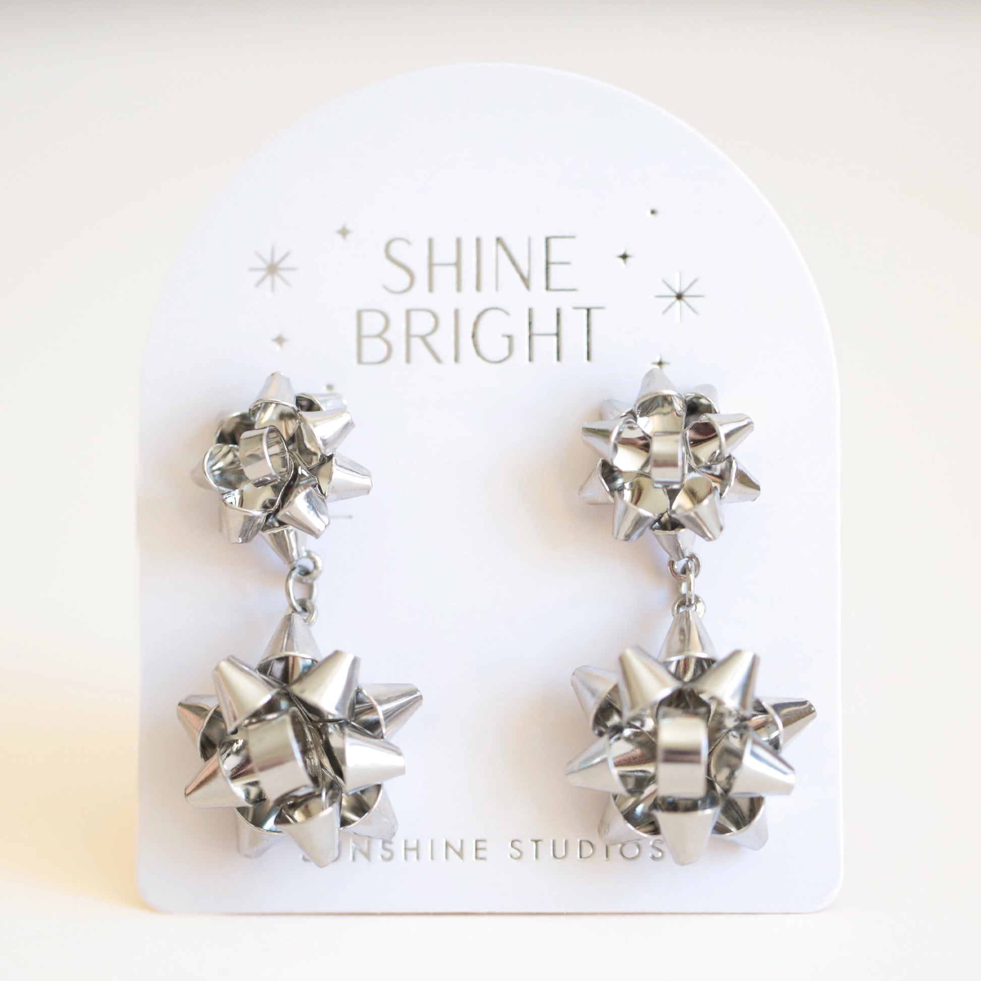 On an ivory background is a pair of silver holiday bow shaped earrings that have a smaller gold bow and a dangling larger bow attached to the bottom. The packaging reads, "Shine Bright Sunshine Studios".