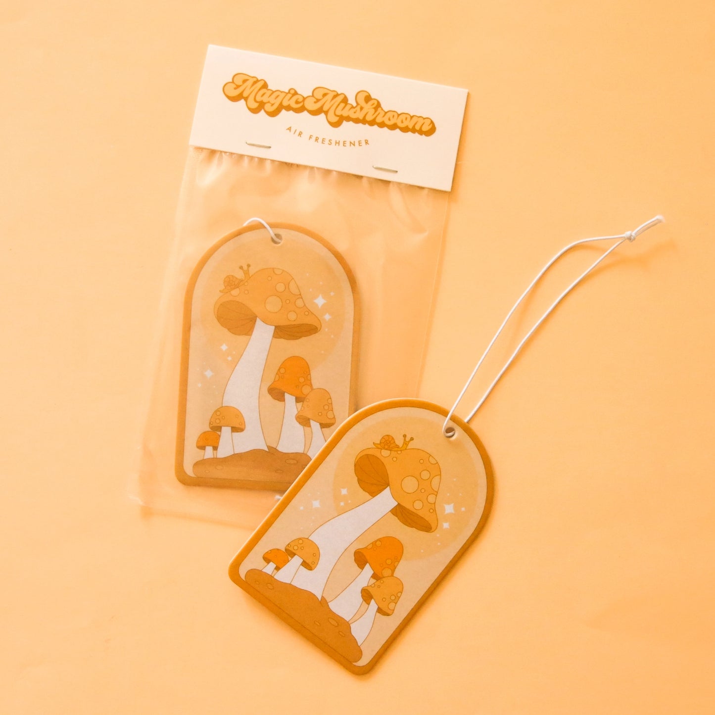 On a light orange background is an arched shaped air freshener with an orange mushroom design and a white elastic loop for hanging. 