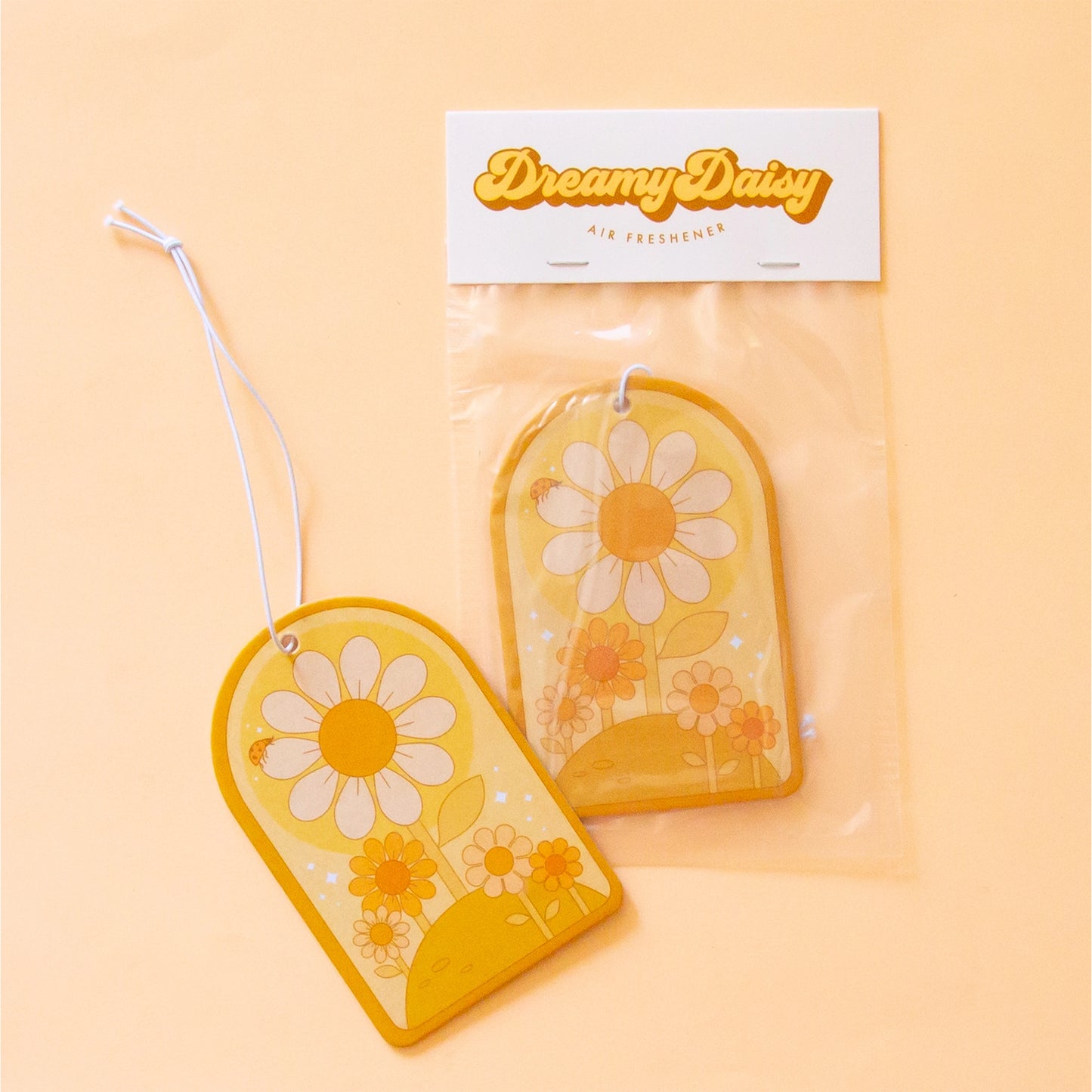 On a peachy background is an arched shaped air freshener with daisy designs and a white elastic loop for hanging. 