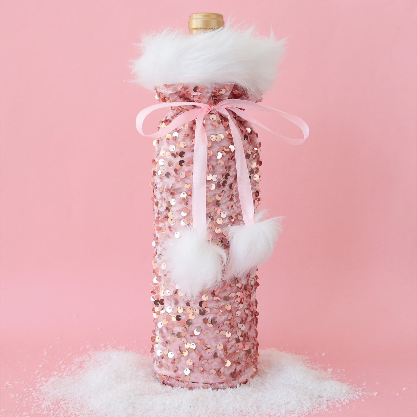 On a pink background is a pink sequin wine gift bag with fuzzy white details and a pom pom pink bow.