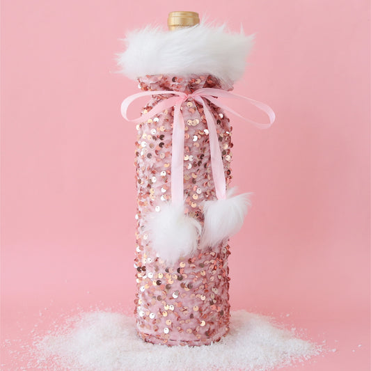 On a pink background is a pink sequin wine gift bag with fuzzy white details and a pom pom pink bow.