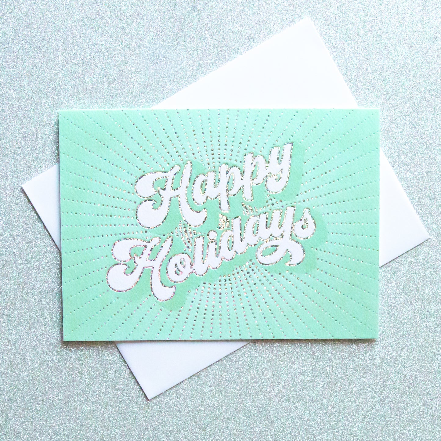 On an ivory background is a teal holiday greeting card with white text that reads, "Happy Holidays" along with a coordinating white envelope. 