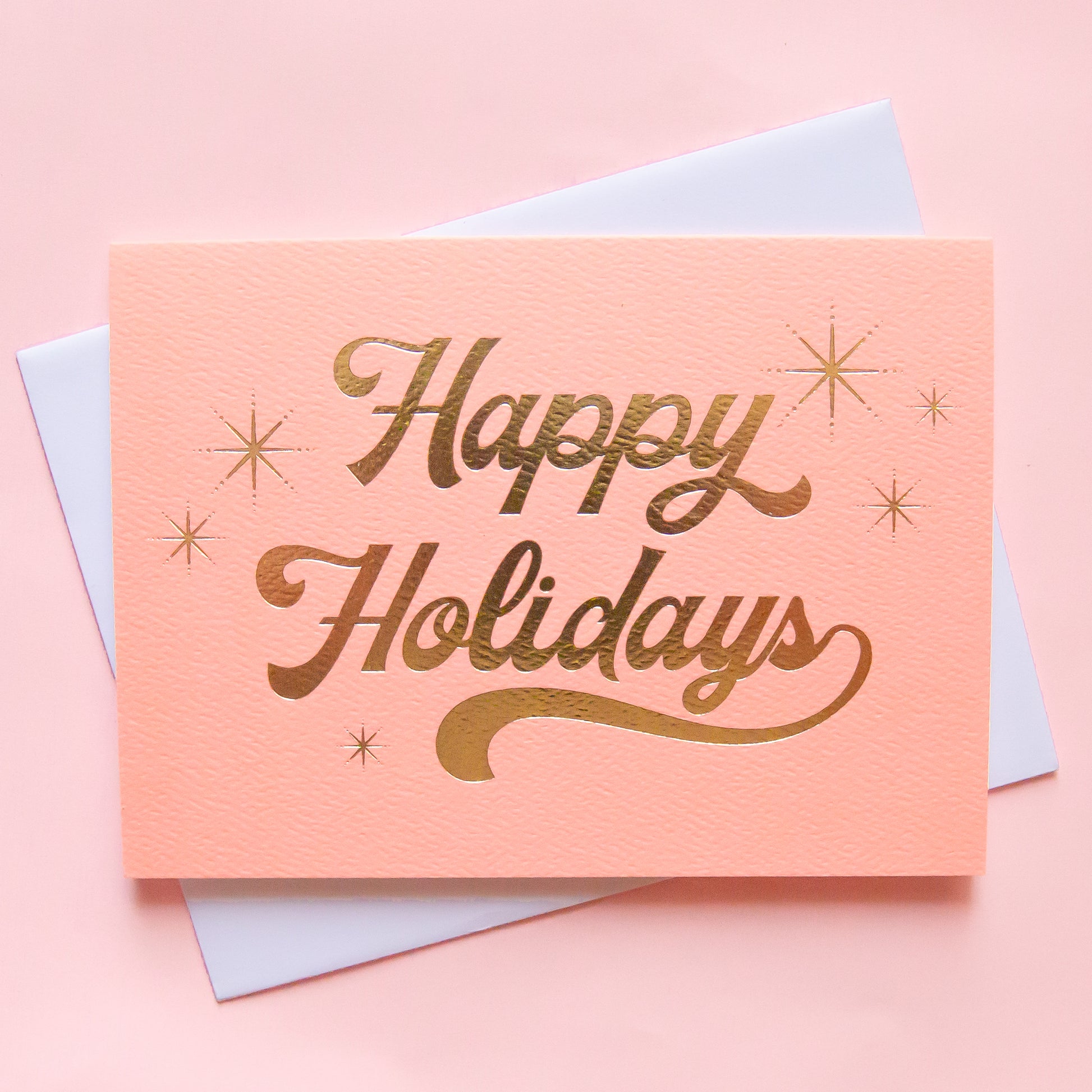 
A peach colored greeting card with cursive text in gold foil that reads happy holidays surrounded by six gold foil starbursts. The card is accompanied by a solid white envelope. 