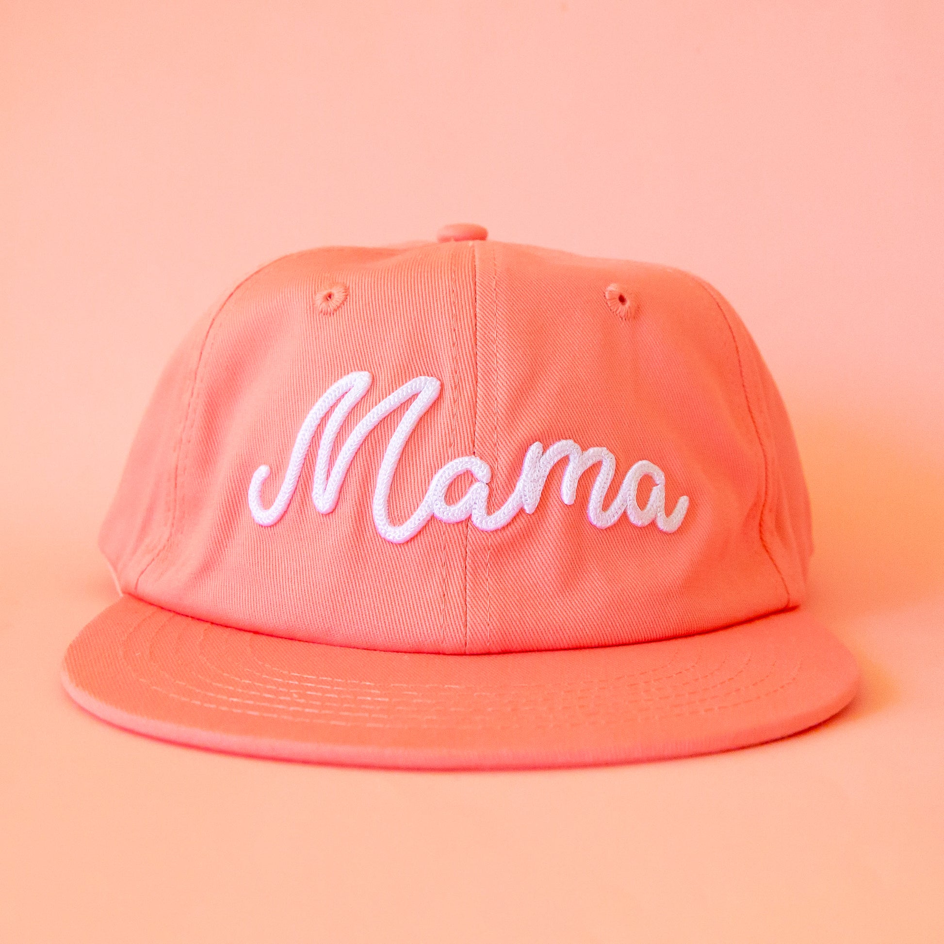 A peachy pink flat brim hat with white embroidered text that reads, 'Mama'.