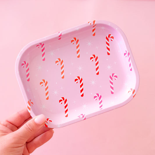 A hand comes in the bottom left corner of frame holding a metal tin tray.  The tray has pink, orange and red candy canes on it with white sparkle detail.  The background of the image is a soft pink color.