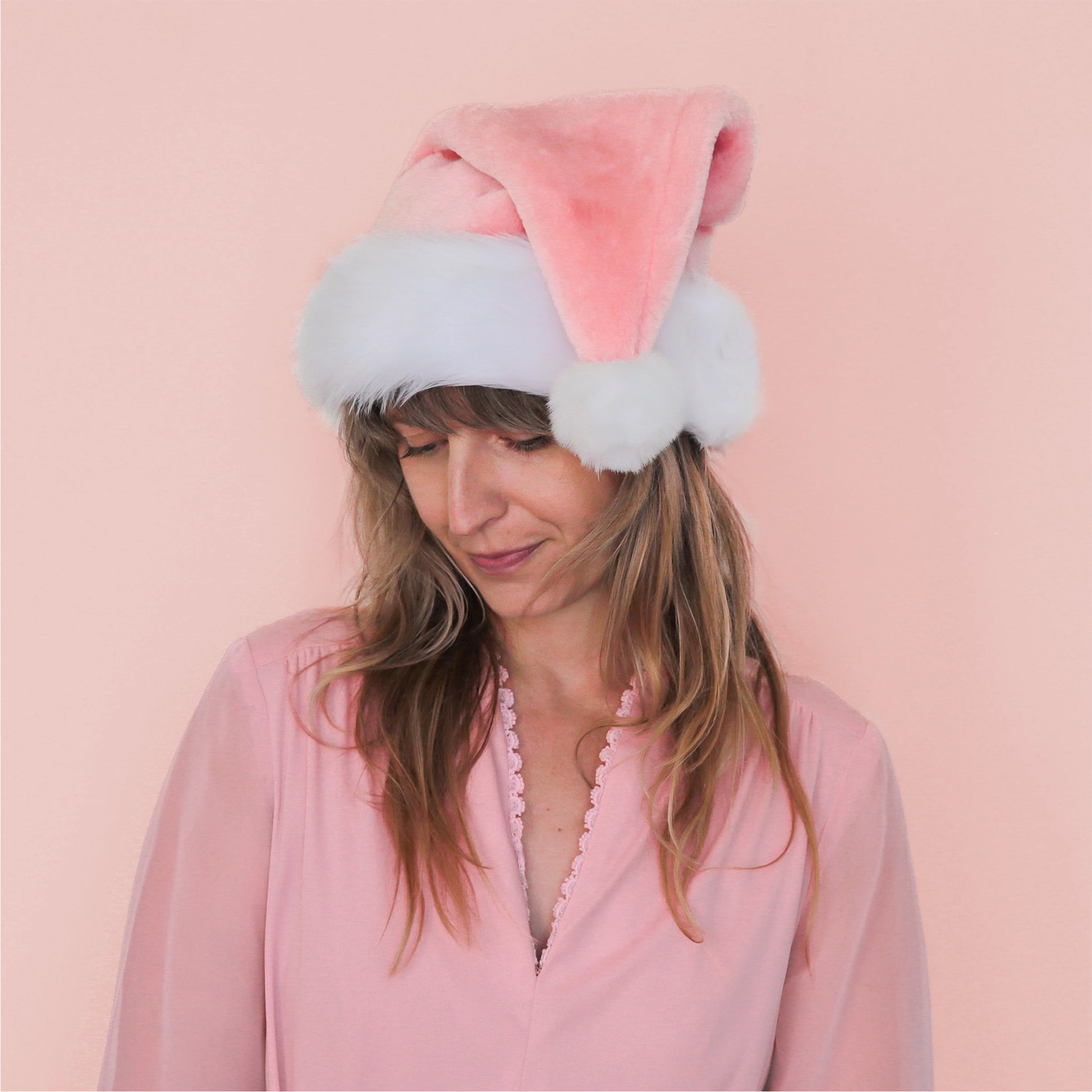 On a pink background is model wearing a pink santa hat with white faux fur detailing.