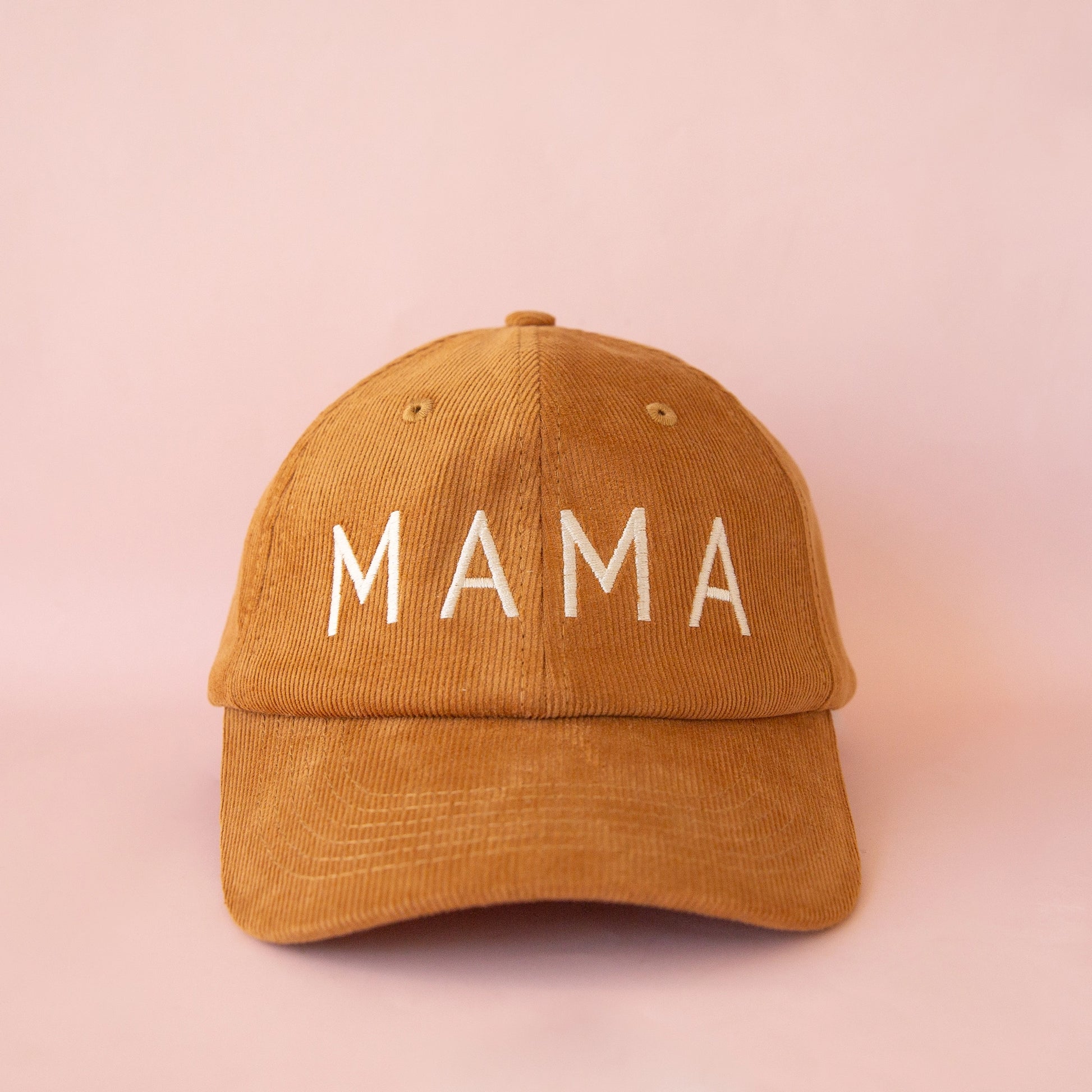 On a pink background is a toffee colored corduroy baseball hat with white embroidered text on the front that reads, "MAMA". 
