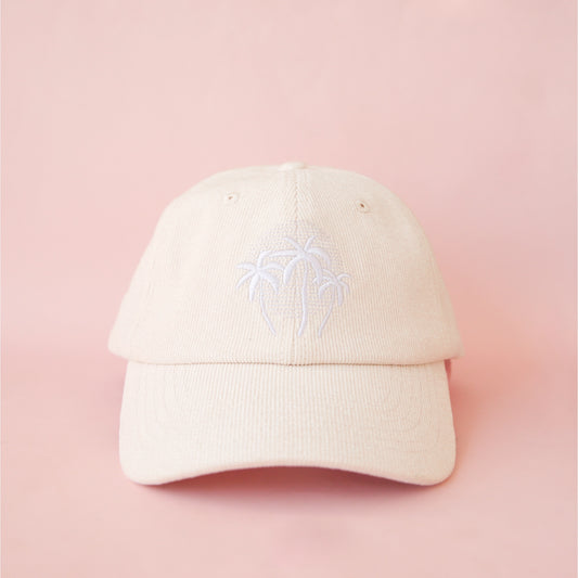 On a pink background is an ivory corduroy hat with a white embroidered palm tree graphic. 