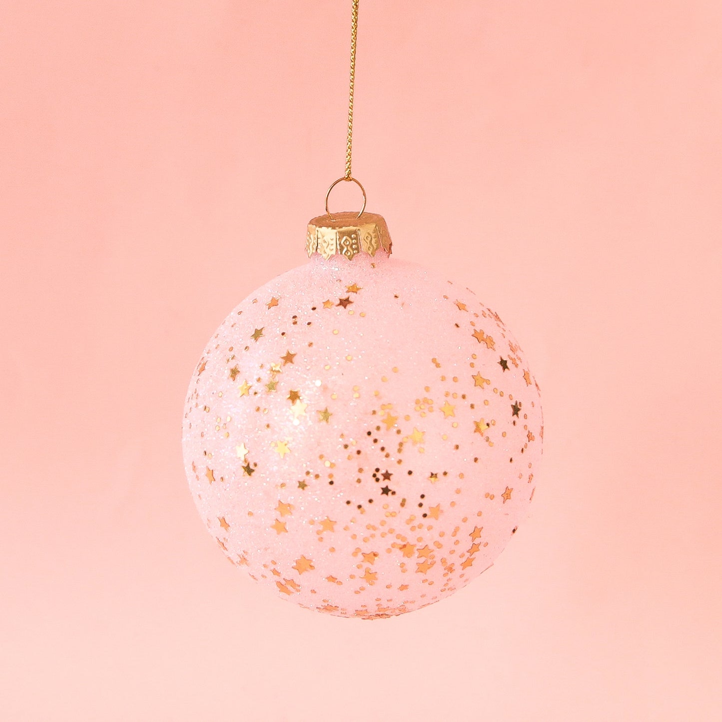 On a peachy background is a light pink ball ornament with small gold stars all over. 