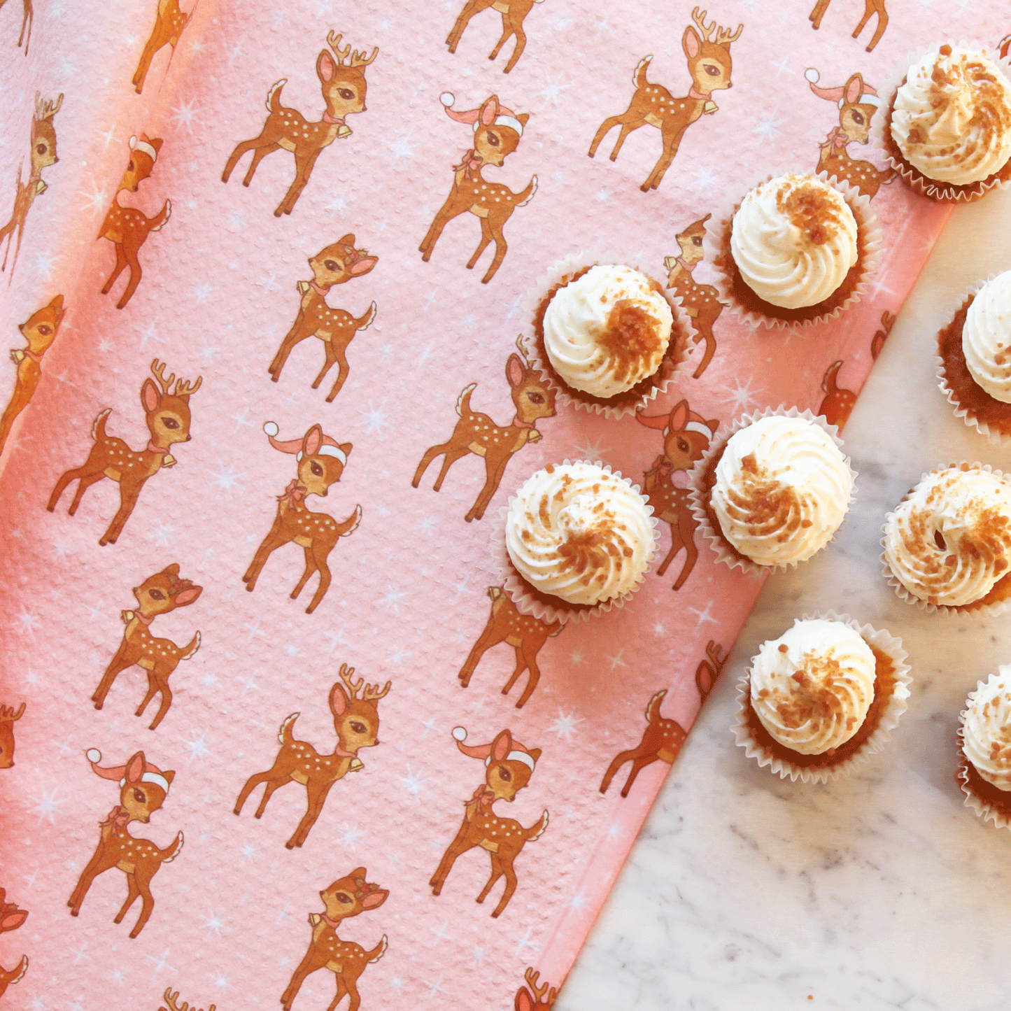 A pink kitchen towel with a brown retro deer pattern and staged next to white frosted cupcakes. 