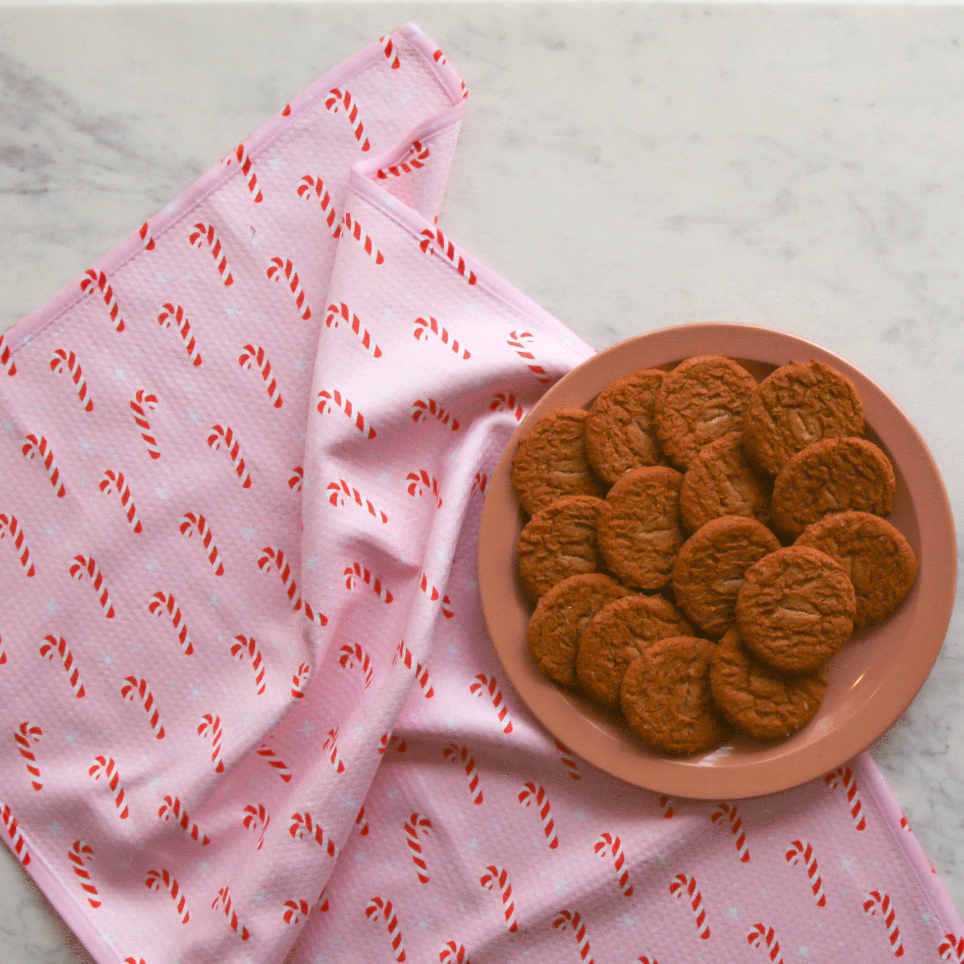 A pink kitchen towel with a red and white candy cane design on a counter next to a plate of gingersnap cookies. 