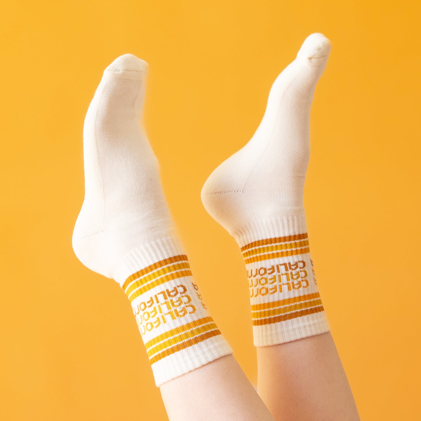 On a peach background is a white socks with rust, yellow and orange stripes and "california" printed three times on the ankle.'