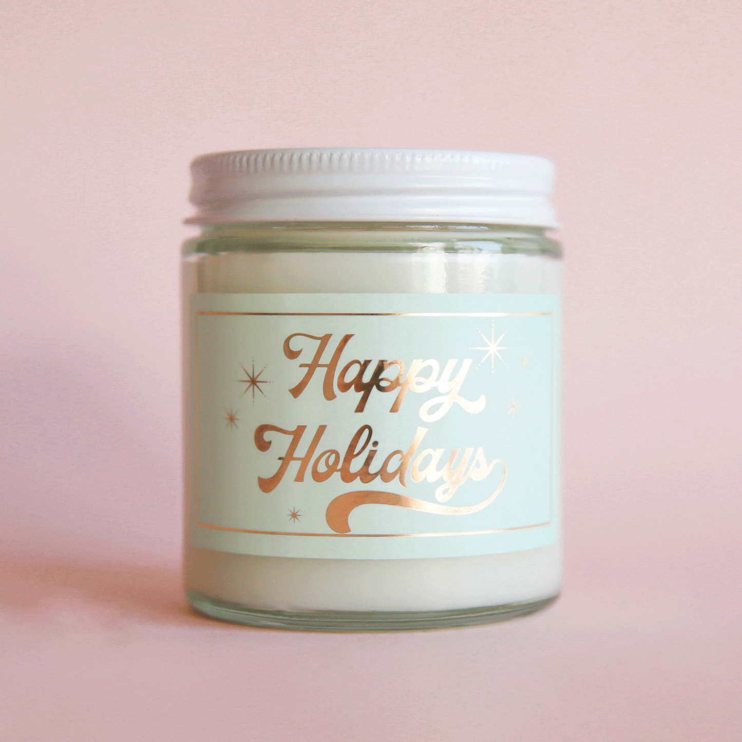 On a pink background is a clear glass candle with a light mint green label and gold text that reads, "Happy Holidays".