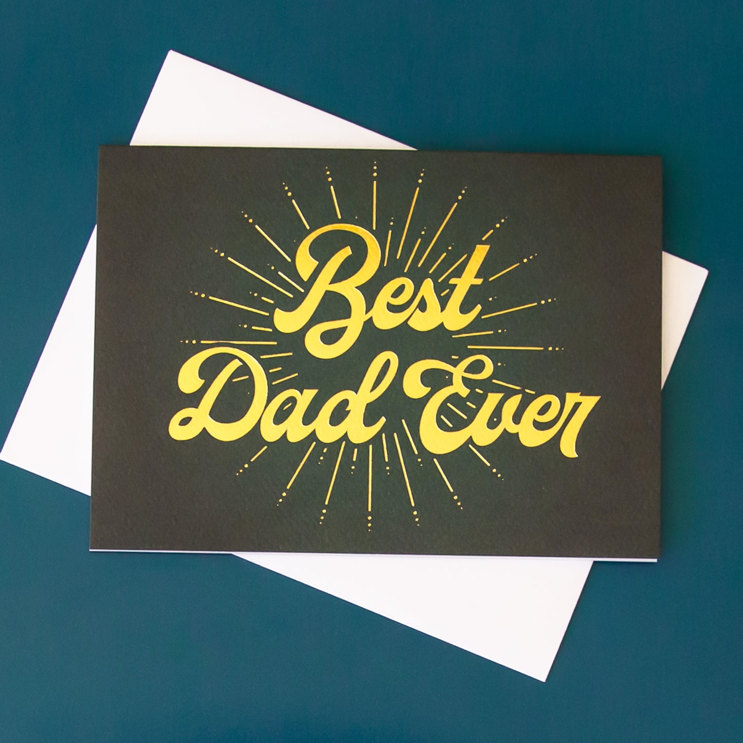 On a light green background is a dark green Father's Day card with gold text across the front that reads, "Best Dad Ever".