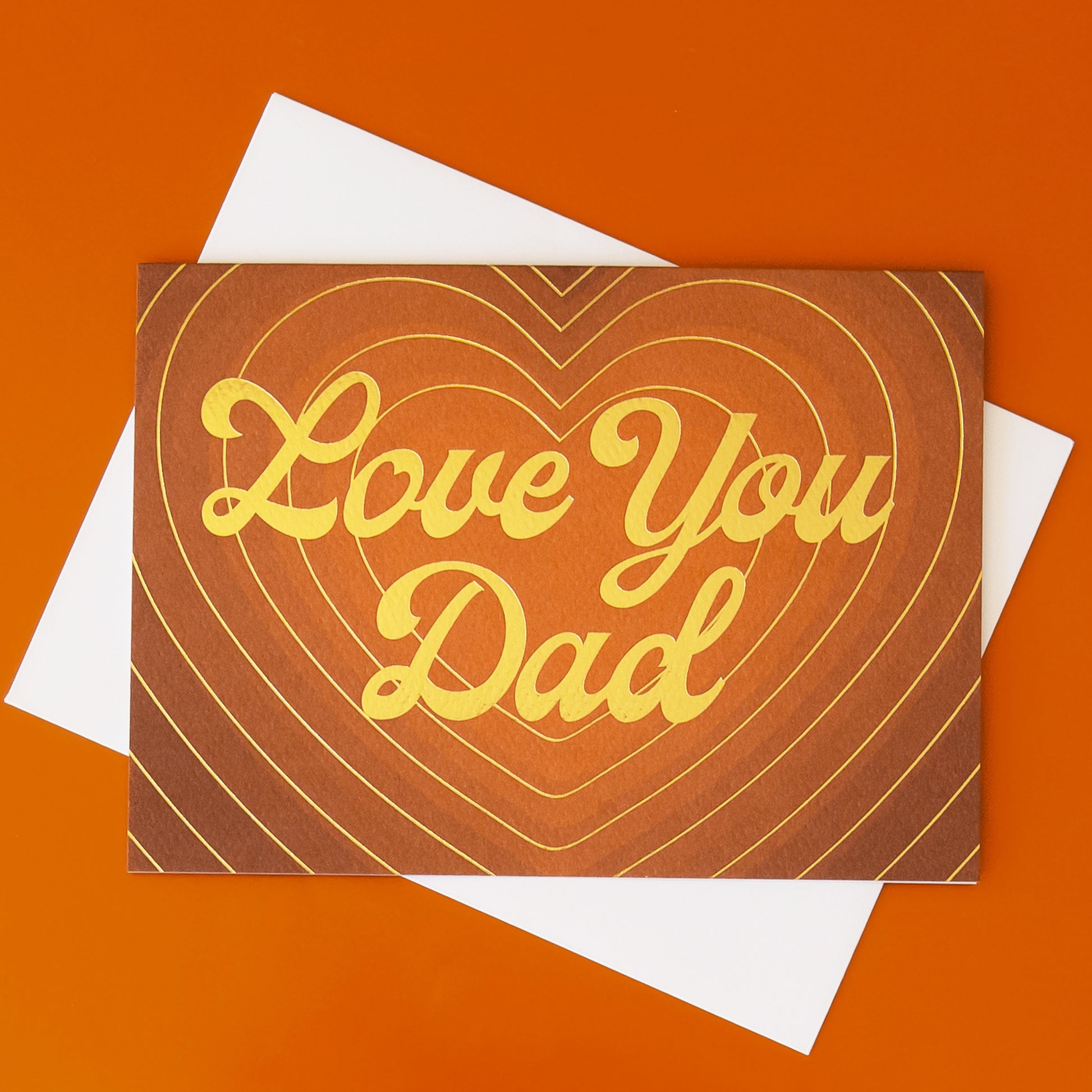 On a light brown background is a brown card with a radiating heart card and gold foiled text that reads, "Love You Dad".