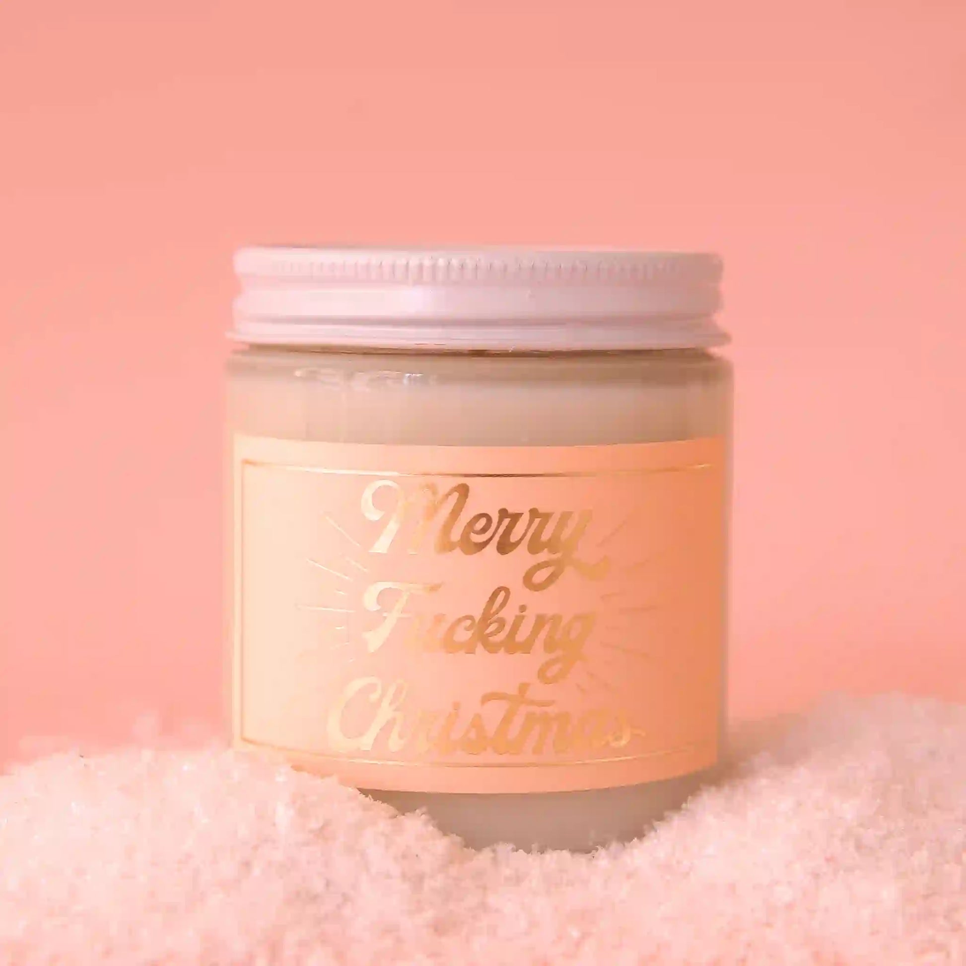 On a pink background is a clear glass jarred candle with a white lid and a peachy label that has gold text reading, "Merry Fucking Christmas".
