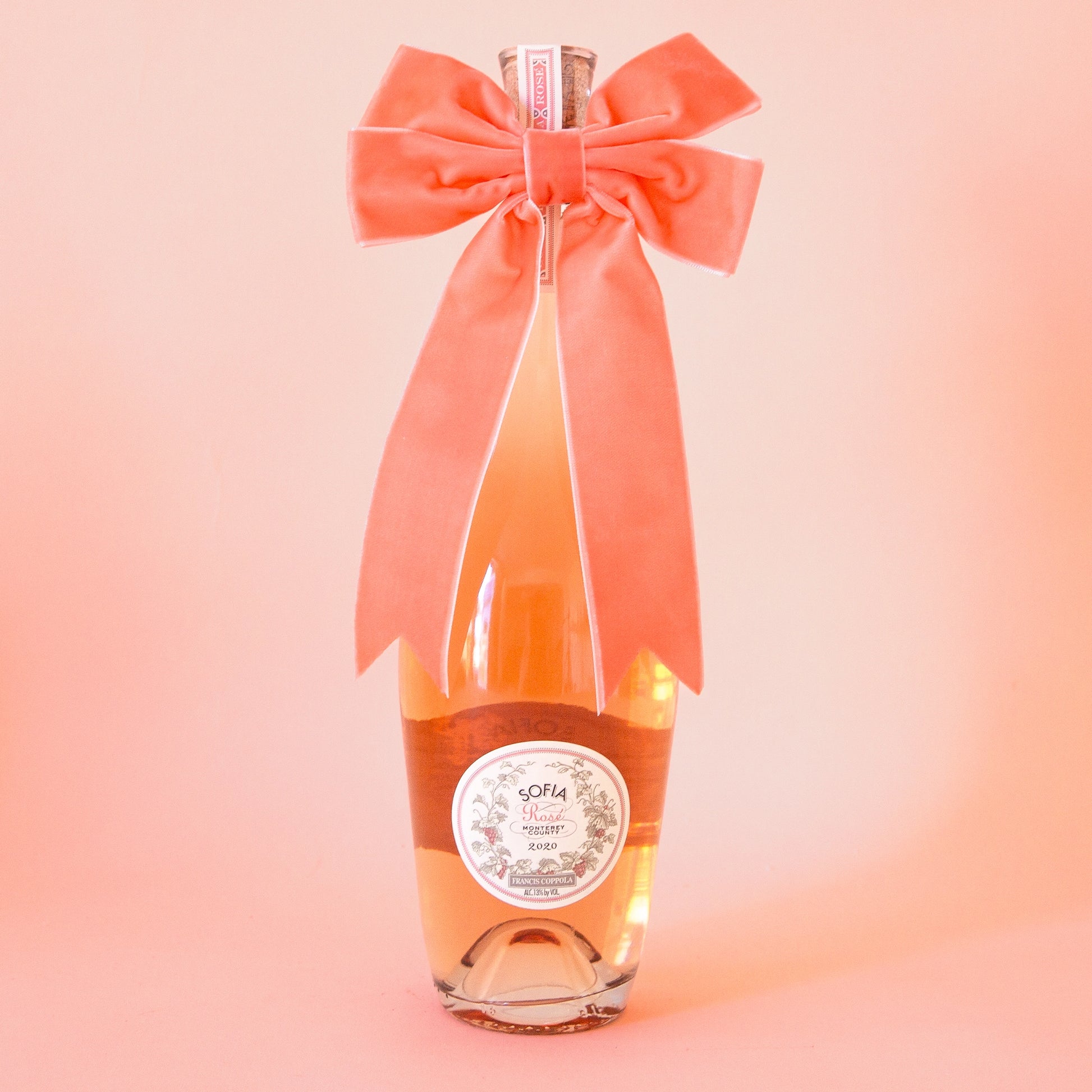 On a peach background is a wine bottle with a peach velvet bow on the front. Wine bottle not included.
