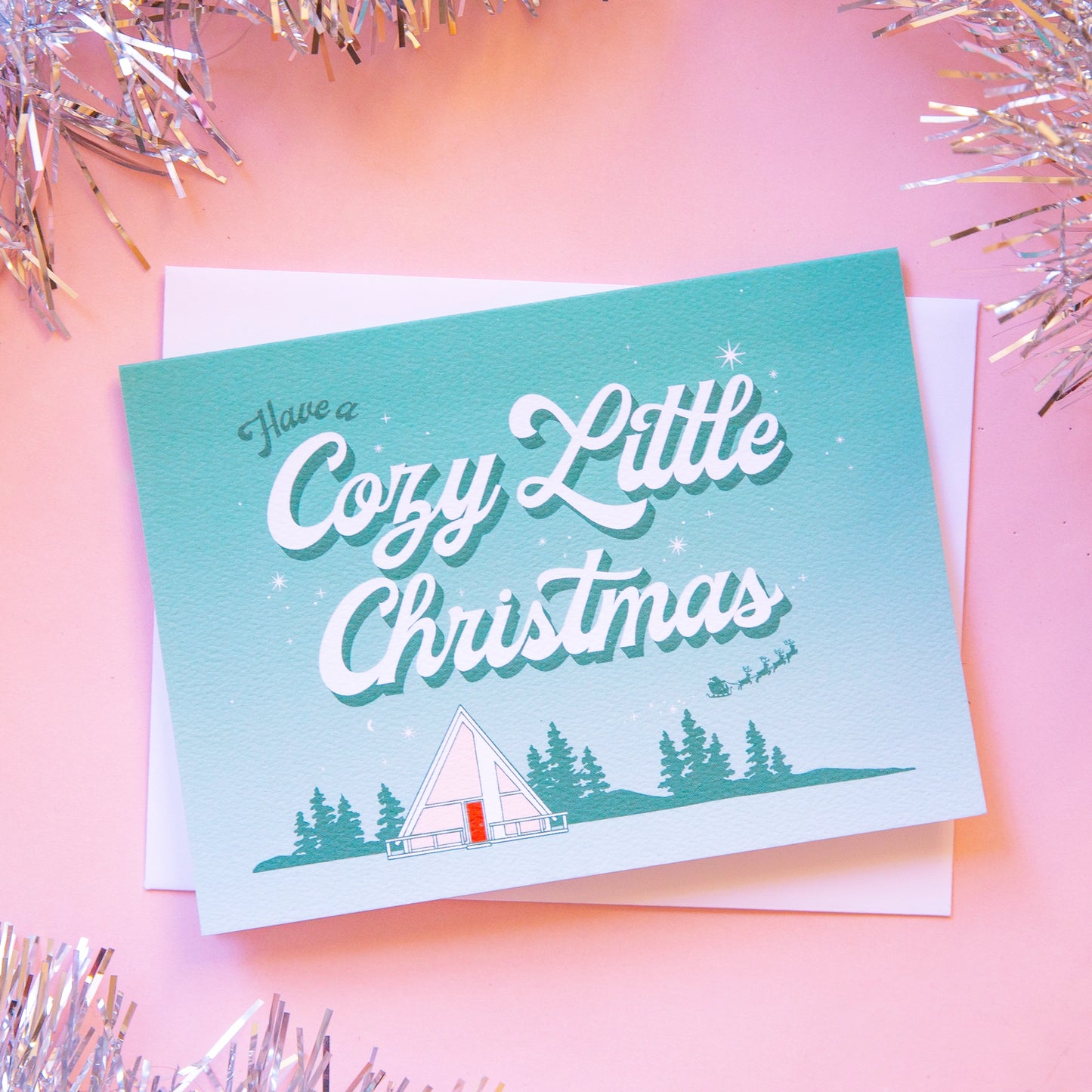 Aqua and white ombre greeting card that reads 'have a cozy little Christmas'. Below the text is a small white tent in a forest scene. A small Santa sleigh and reindeer fly across the sky. The card is accompanied by a white envelope.