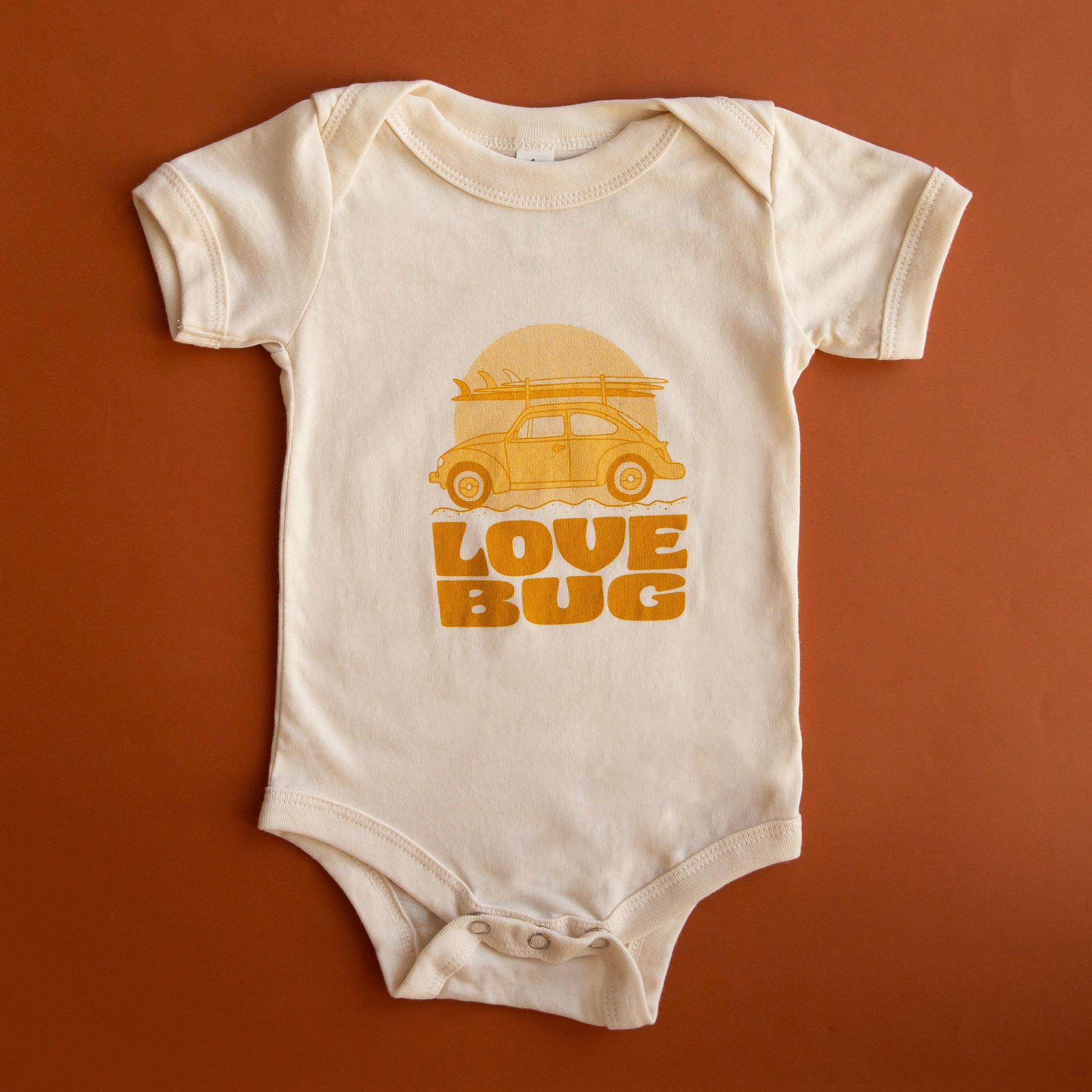 On a burnt orange background is an ivory onesie with a yellow orange VW bug graphic with surfboards on top along with text below that reads, "Love Bug".