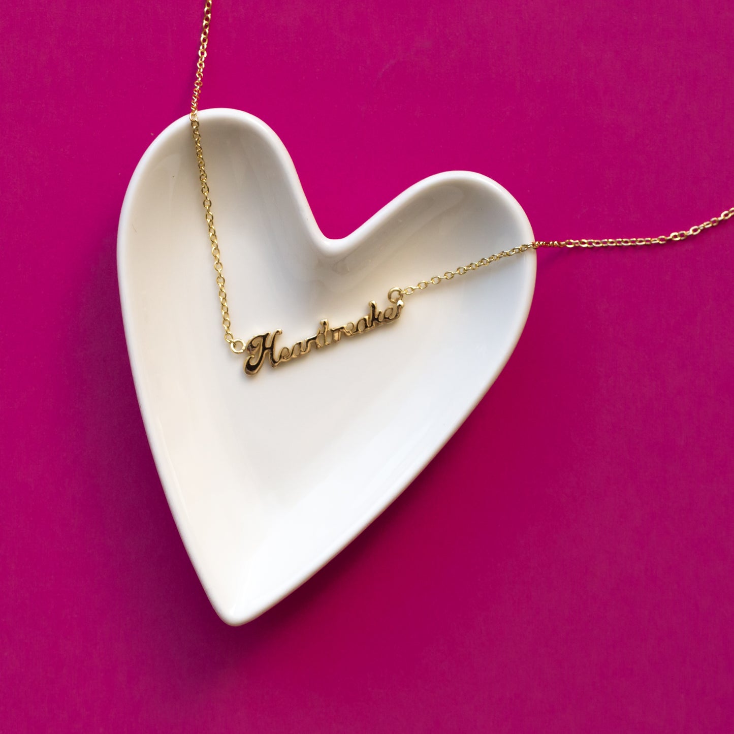 A white, ceramic, heart shaped trinket dish sits on a hot pink background.  A gold nameplate style necklace that reads, "heartbreaker" lies on the heart dish.