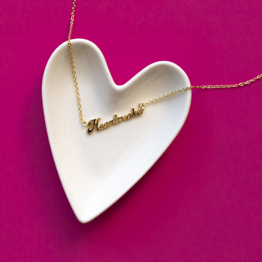 A white, ceramic, heart shaped trinket dish sits on a hot pink background.  A gold nameplate style necklace that reads, "heartbreaker" lies on the heart dish.