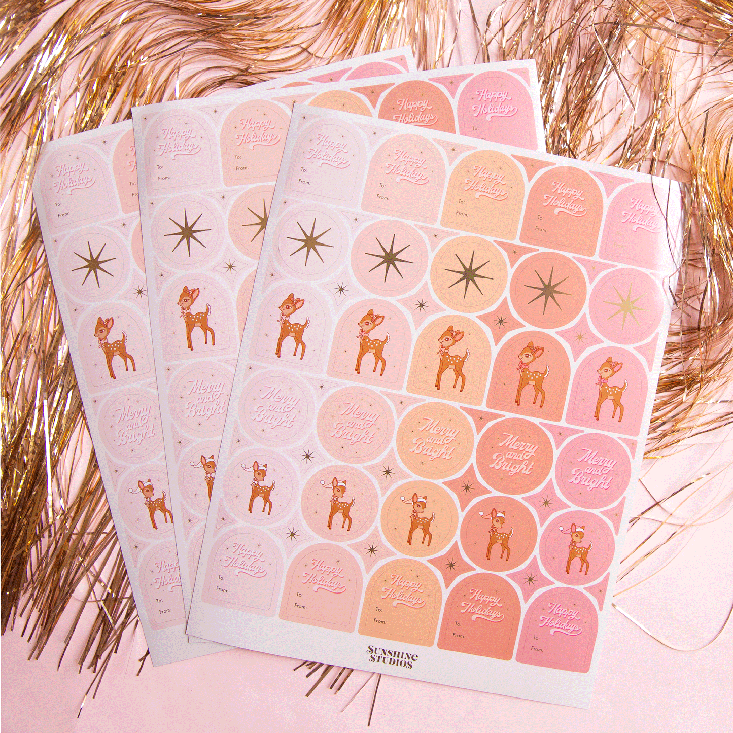 On a pink background is three sheets of gift label stickers with shapes of circles and arches along with text that reads, "Happy Holidays", a gold star, or a retro deer graphic.