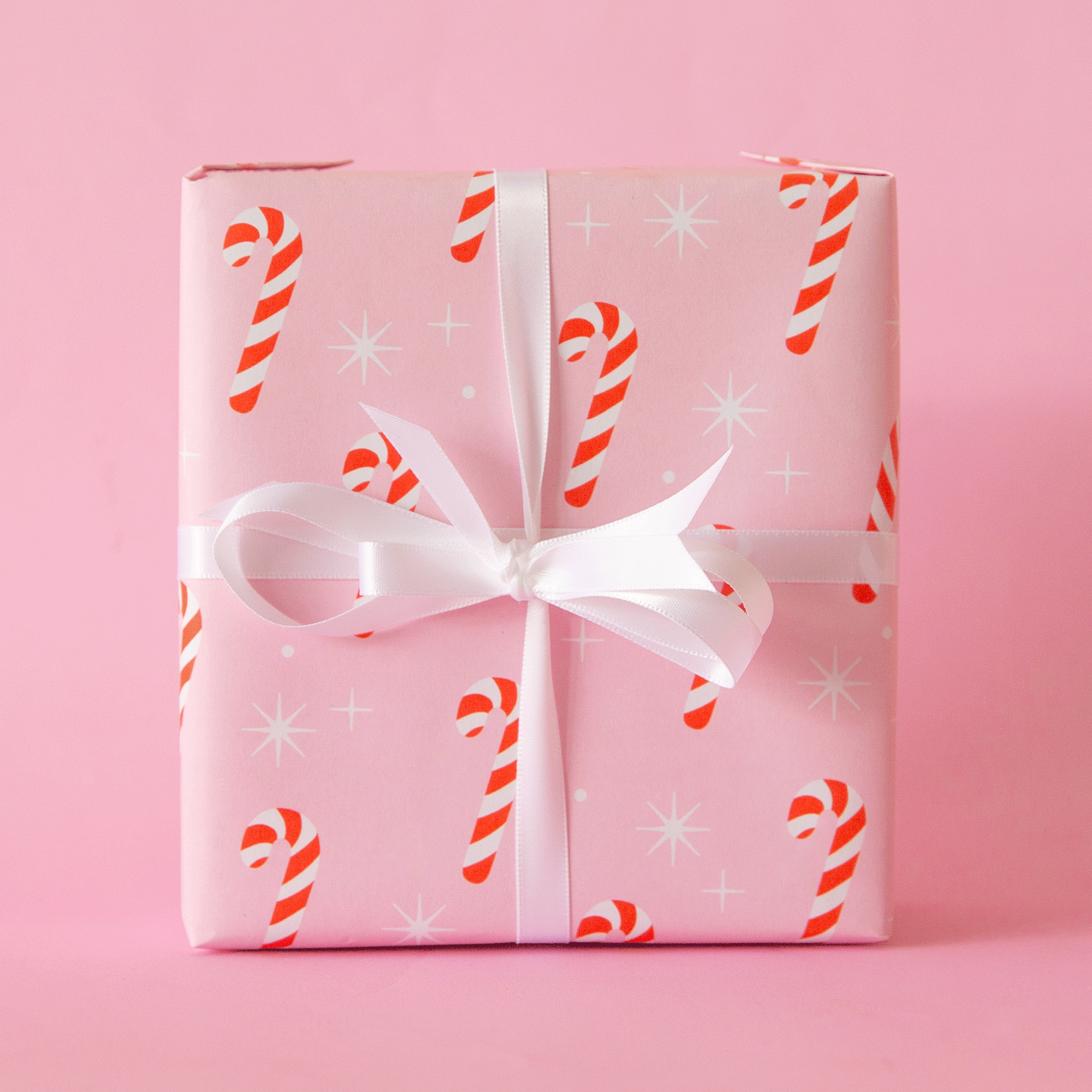 A single sheet of gift wrap with a blush background and a repeating red and white candy cane print. The gift wrap is reversible and the other side features a cool toned pink background with an all over red and white candy cane design but the candy canes are slightly smaller than the reverse.