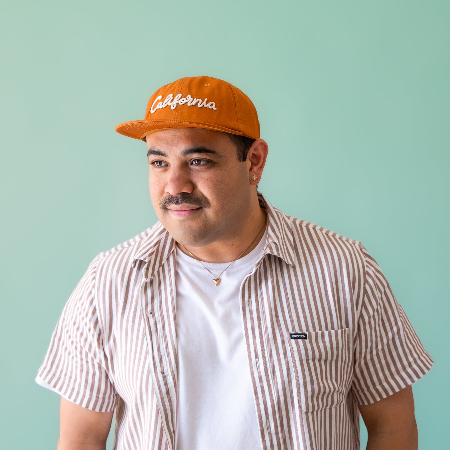 A burnt orange flat brimmed baseball hat with ivory embroidered text that reads, "California" across the front.