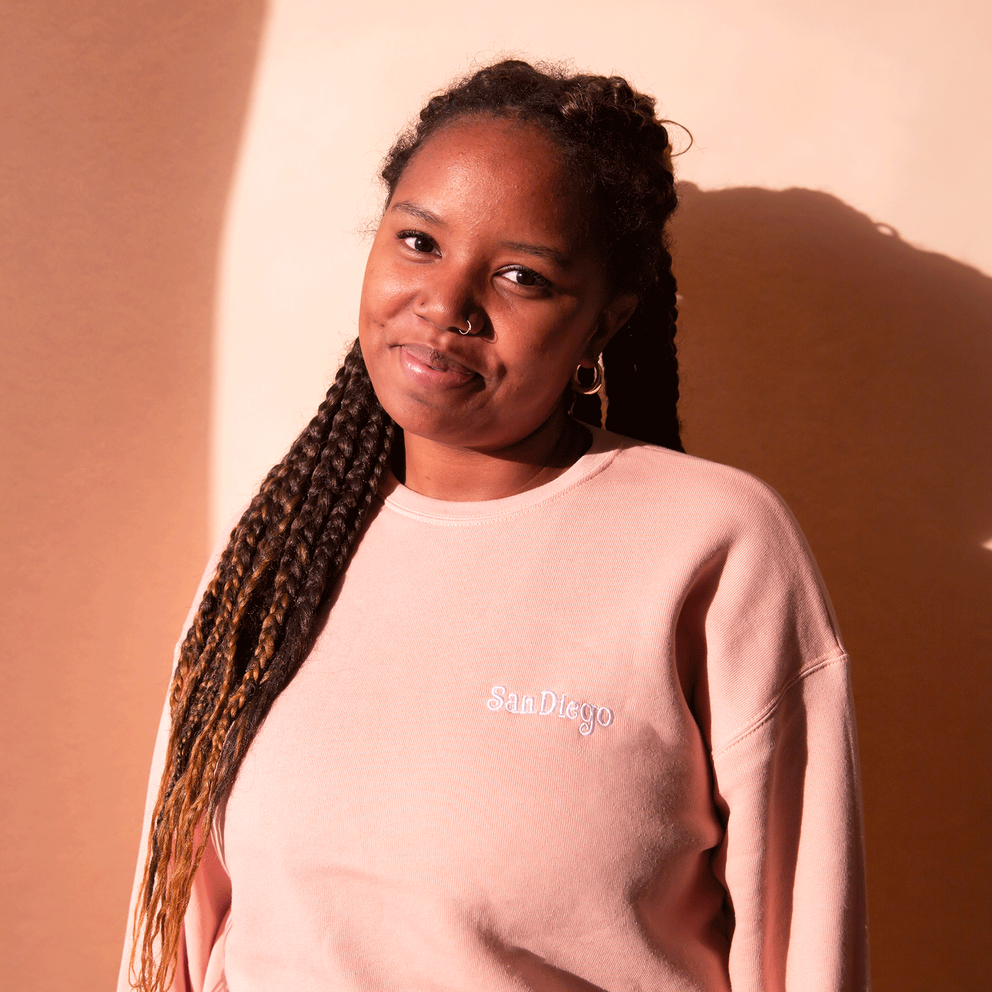 On a peach background is a peachy pink pull over sweatshirt with a white embroidered "San Diego" on the front. 