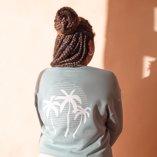 On a peach background is a light blue crewneck sweatshirt with a white palm tree sunset graphic on the back.