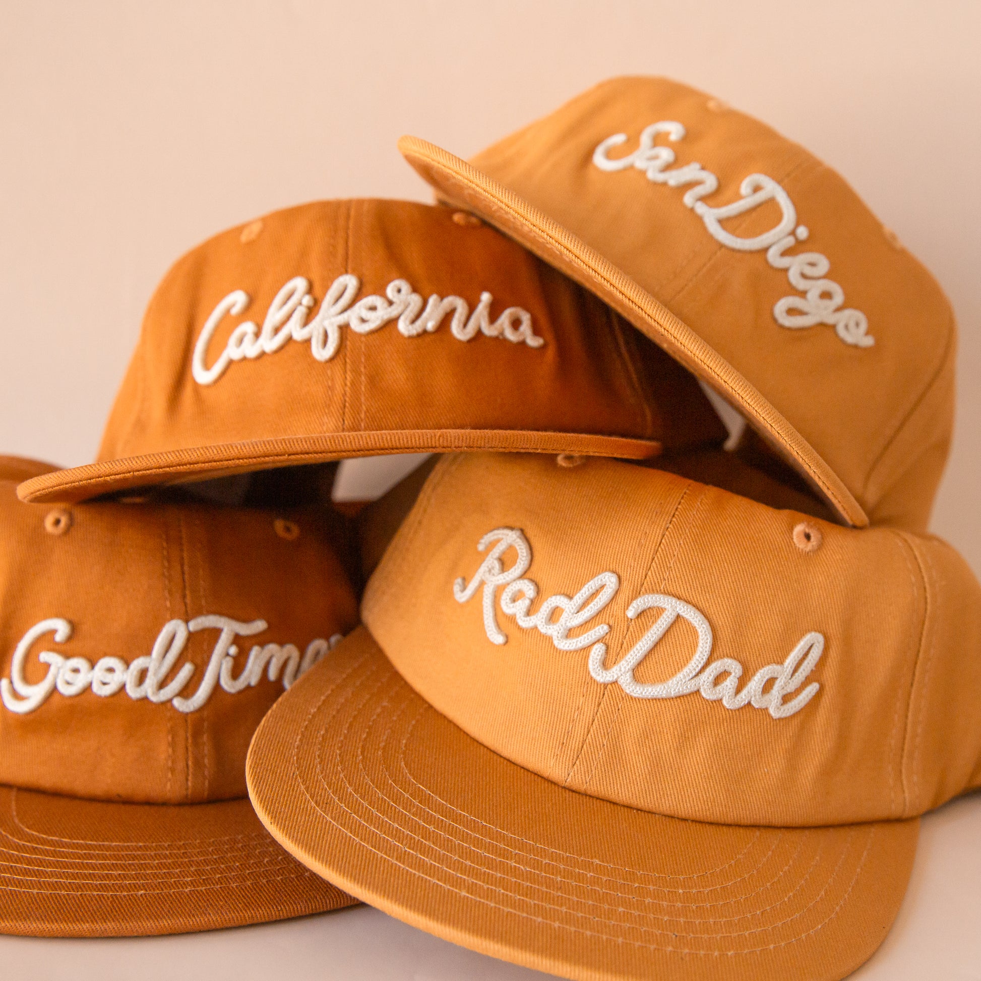 A photograph of the different embroidered phrases on burnt orange flat brimmed hats.