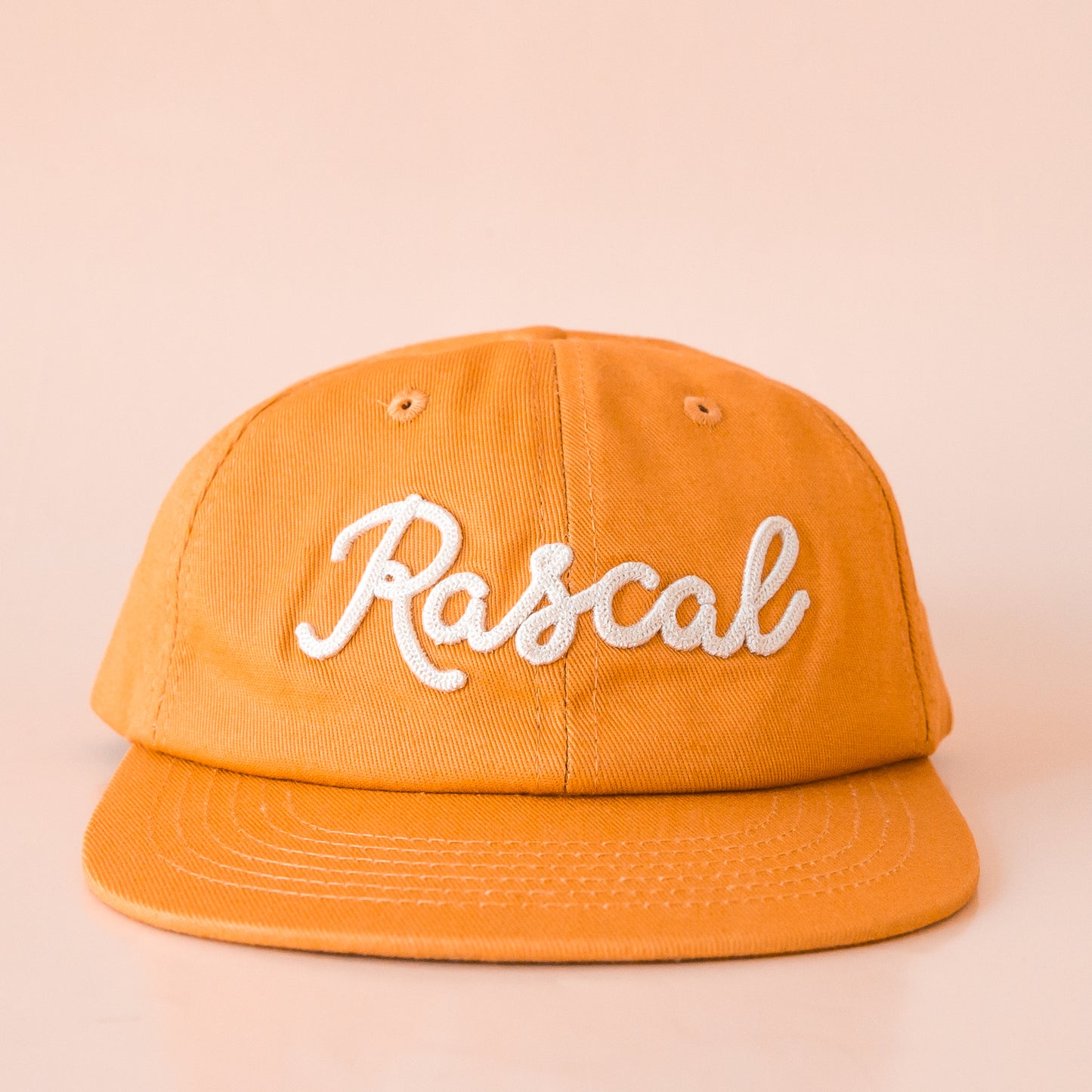 A burnt orange flat brimmed hat with white embroidered text that reads, "Rascal".